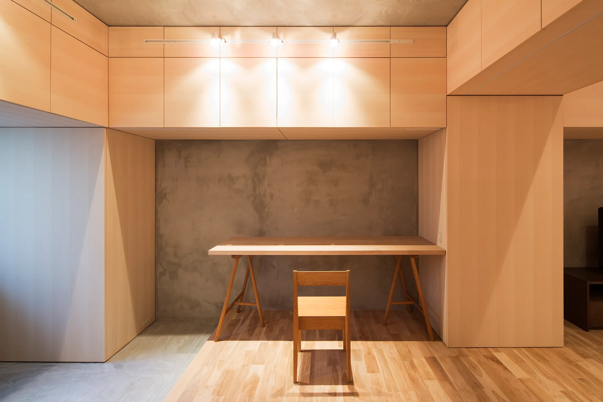 A wooden trestle desk and chair are set against a concrete wall inside an alcove surrounded by blonde wood cabinetry.