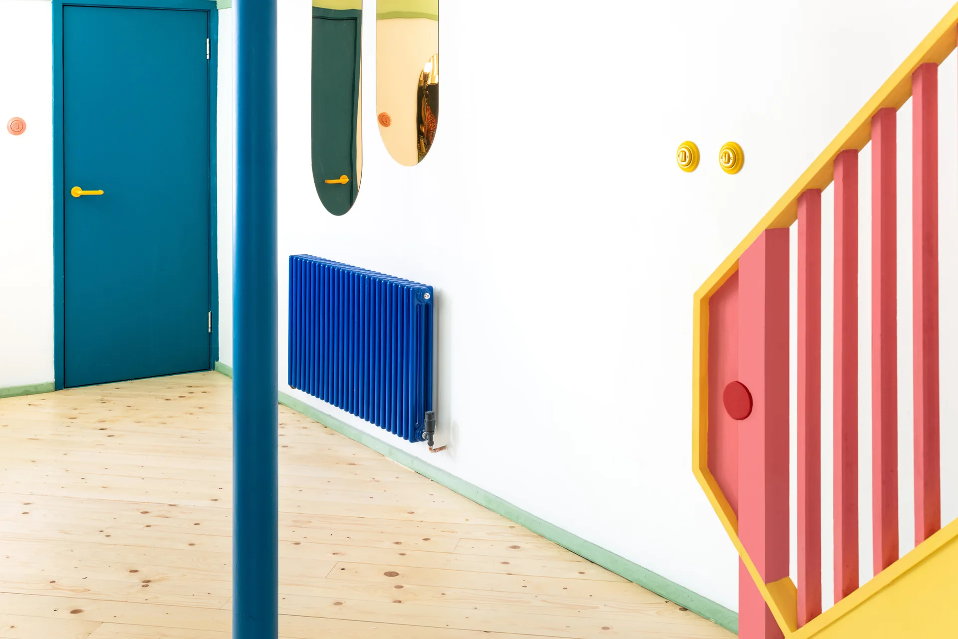 A bright white hallway featuring light wood floors, a blue door, a matching blue radiator, and a red and yellow staircase railing.