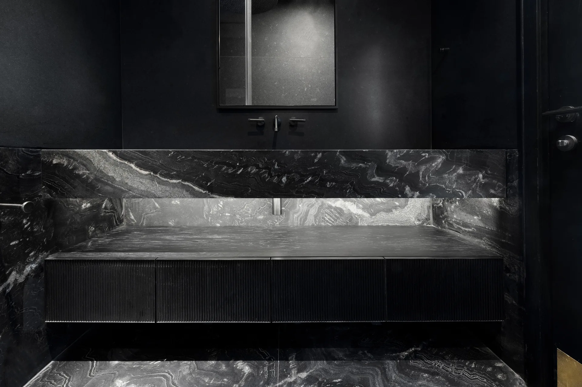 A minimalist bathroom with a black veined stone vanity, fluted dark cabinetry, and integrated lighting beneath a rectangular mirror.