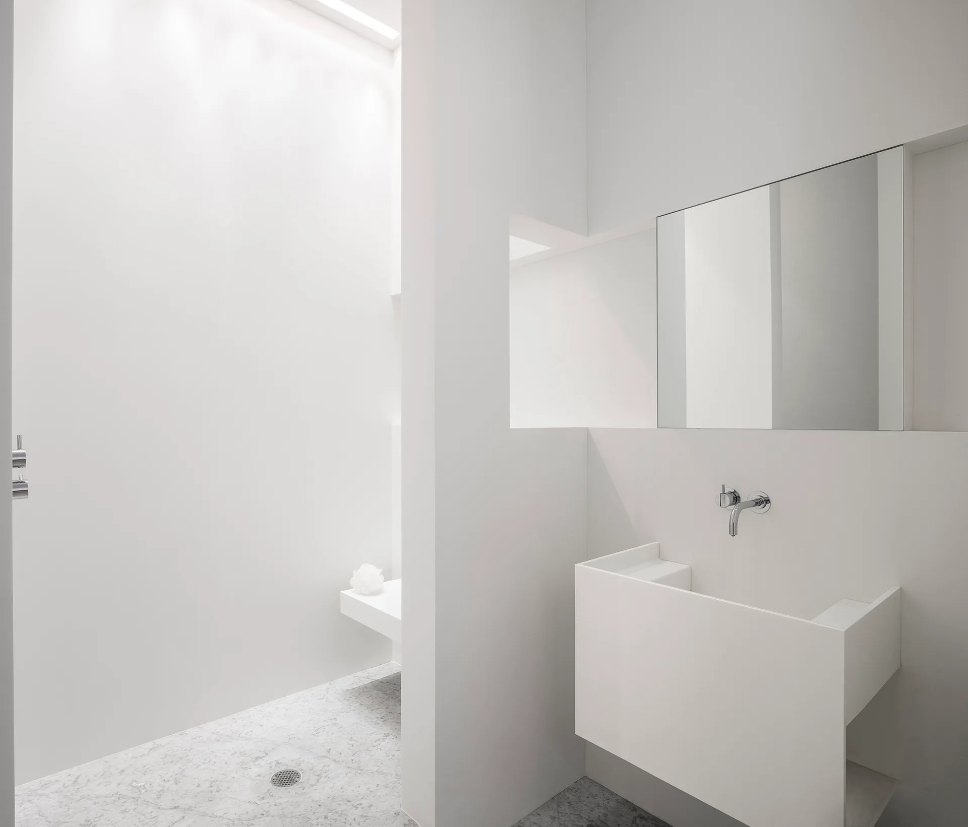 An all-white minimalist bathroom features a floating block vanity, a recessed mirror, and an open shower area with grey veined marble tile flooring.