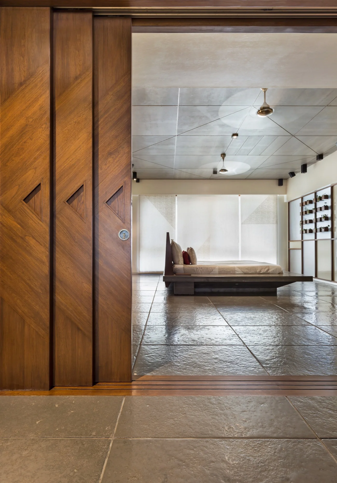 A bedroom with large wooden sliding doors featuring geometric cutouts, a low platform bed, a paneled grey ceiling, and polished stone tile floors.