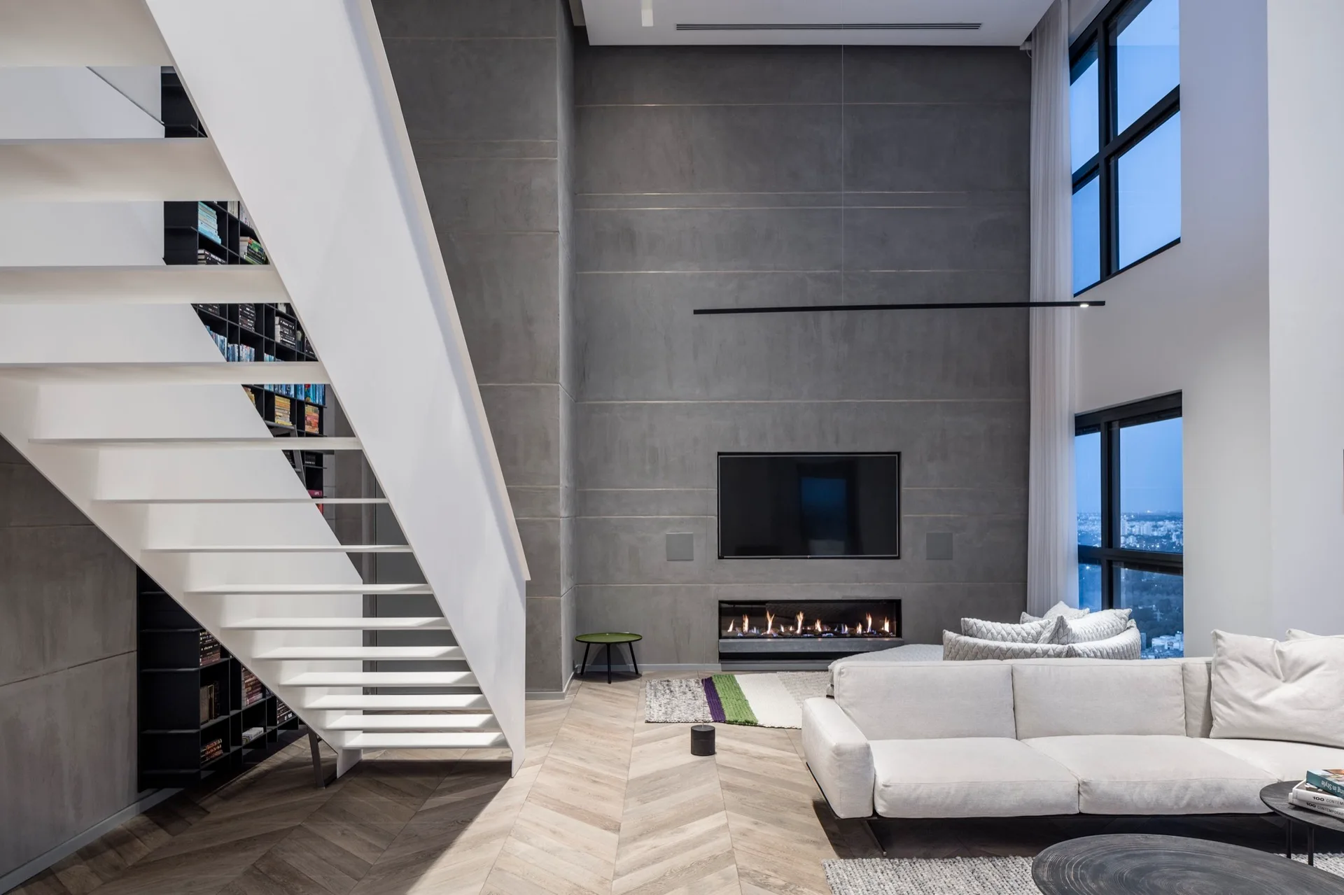 A modern double-height living room featuring a white open-back staircase, a grey paneled fireplace wall, and herringbone flooring.