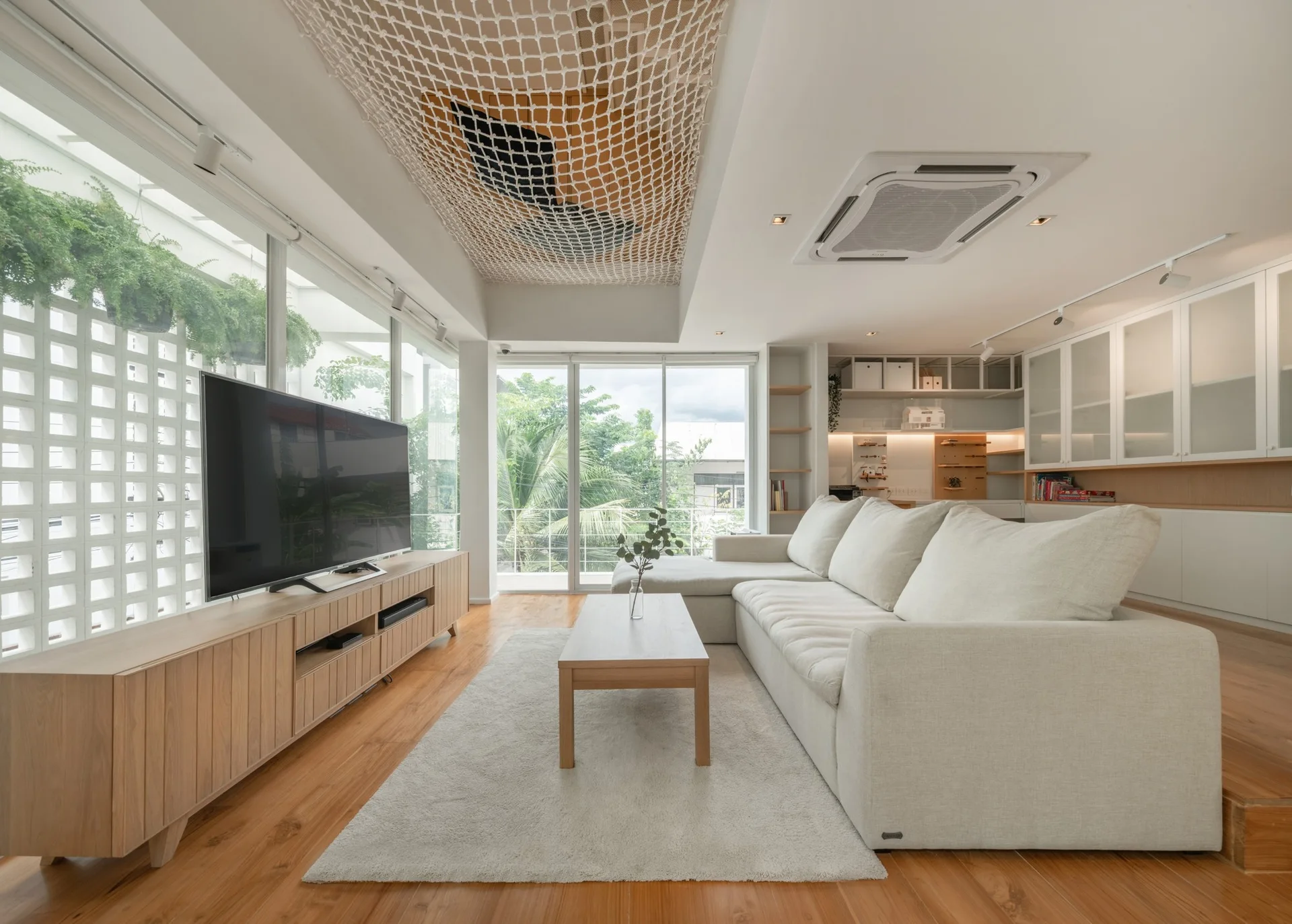 A minimalist living room featuring a light wood media console, white sectional sofa, and a unique woven rope net suspended from the ceiling.