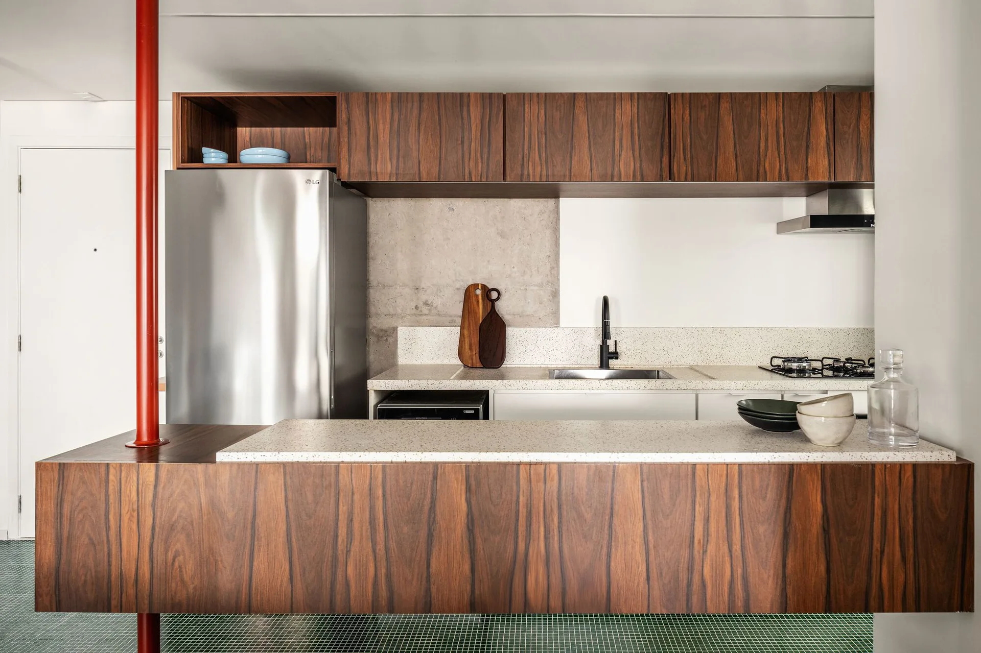 A modern kitchen featuring dark wood cabinets, speckled stone-like countertops, and a prominent red structural column beside a stainless steel refrigerator.