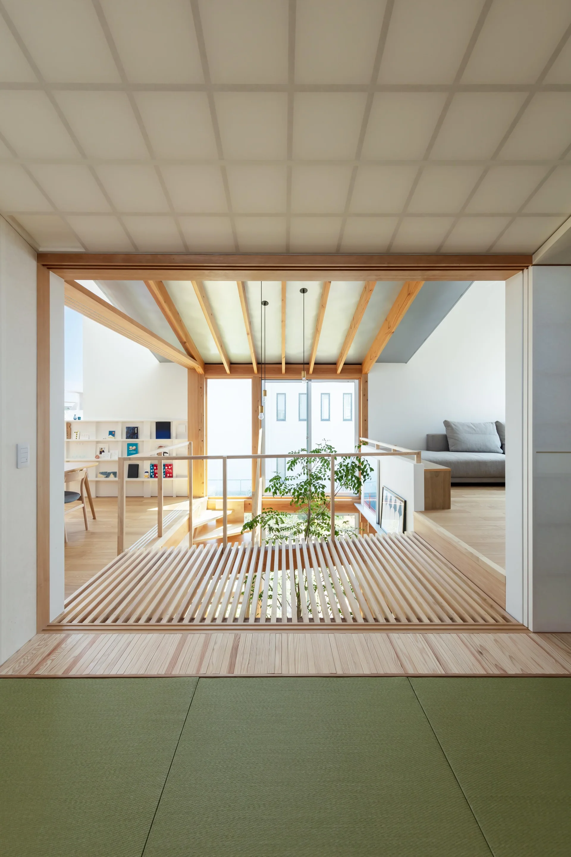 A multi-level minimalist interior featuring green tatami mats in the foreground, wood-slatted flooring over a central void, and an indoor tree.