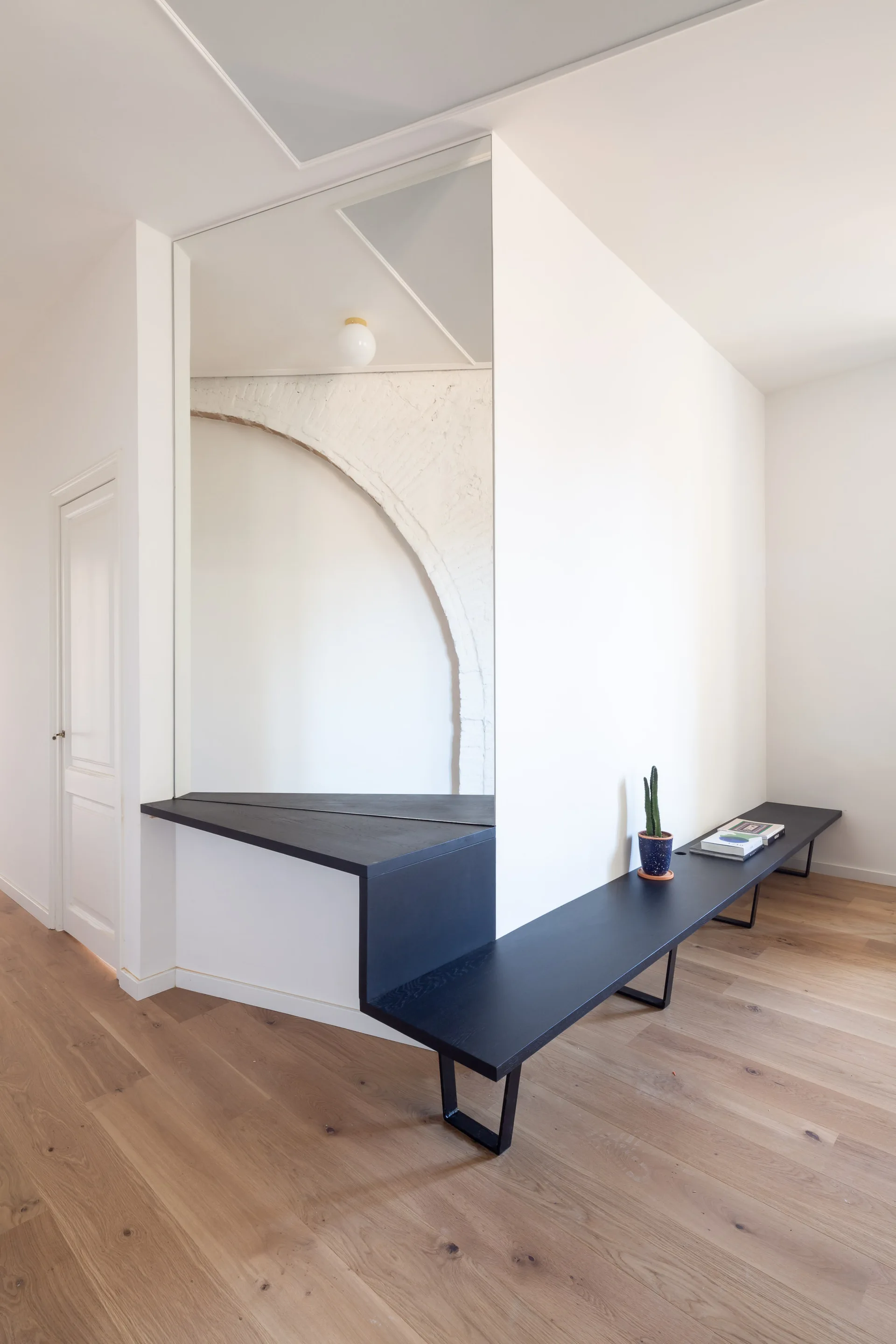 A minimalist hallway features a long custom black wooden bench and a large mirror reflecting a white painted brick arch.