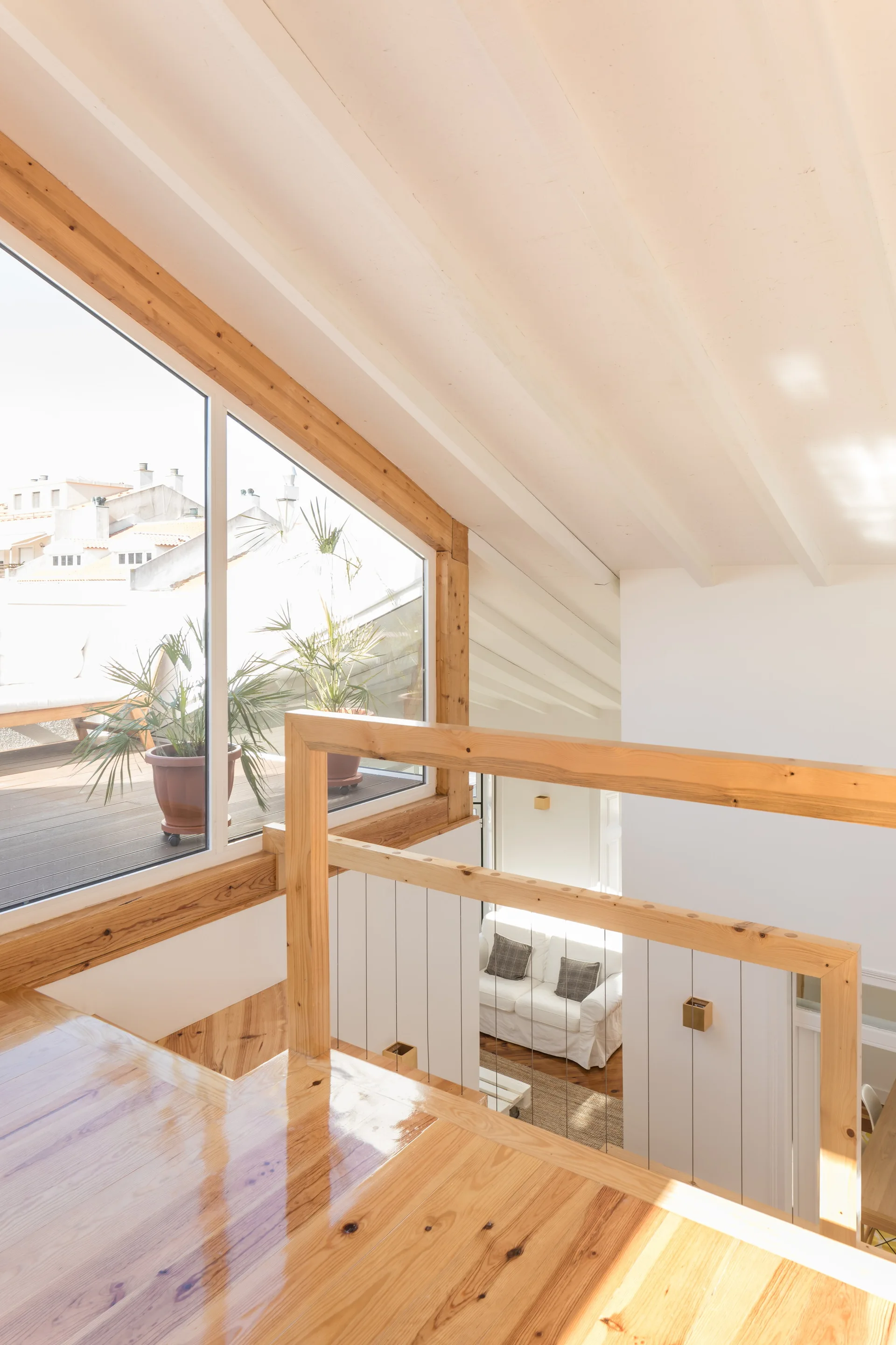 High-angle view of a glossy wooden staircase landing with a timber railing overlooking a sunlit white living room and a plant-filled terrace.