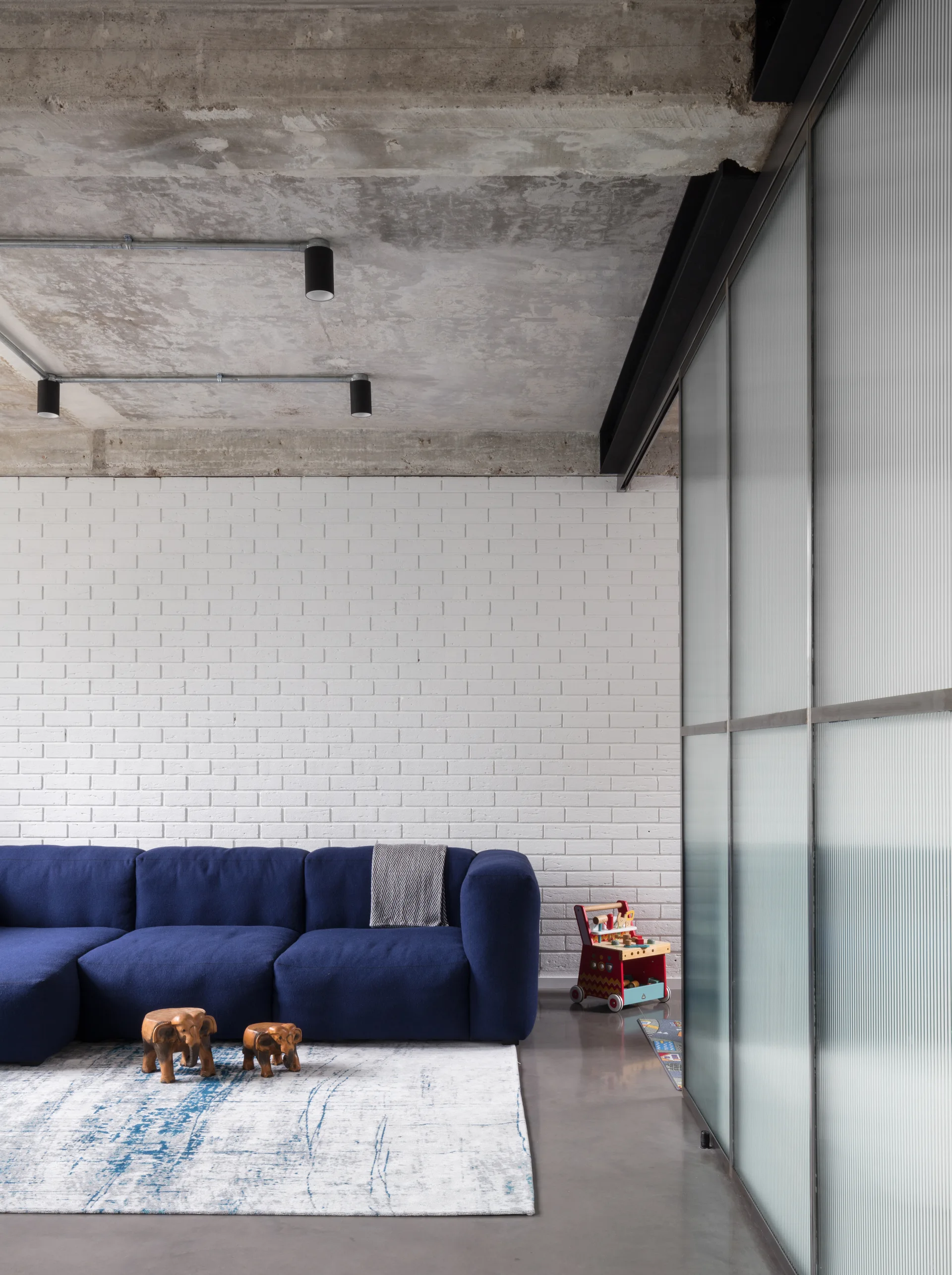 A blue modular sofa sits against a white painted brick wall under an exposed concrete ceiling with a ribbed glass partition on the right.