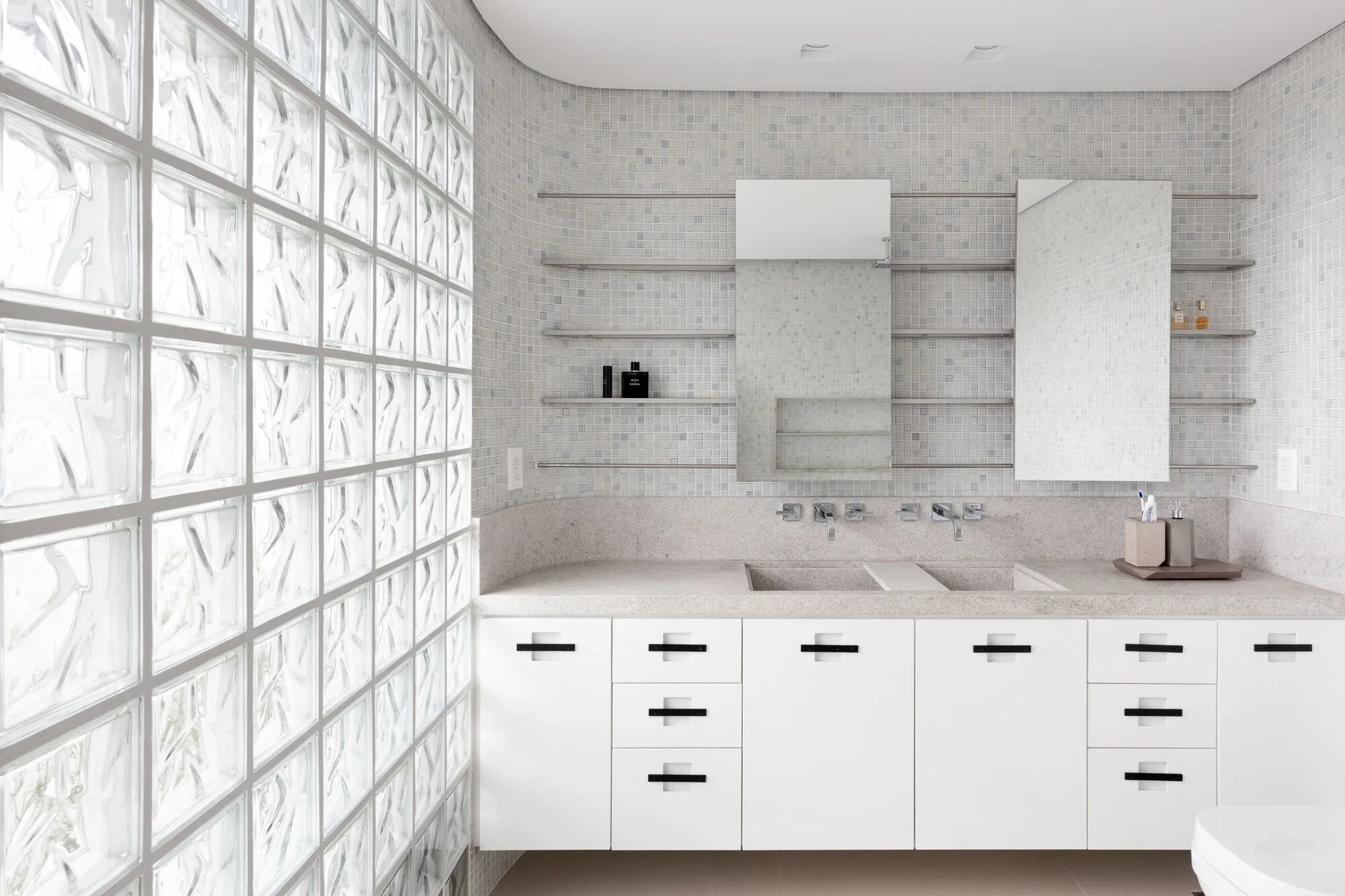 A modern minimalist bathroom featuring a large glass block wall, a white vanity with integrated sinks, and light gray mosaic wall tiles.
