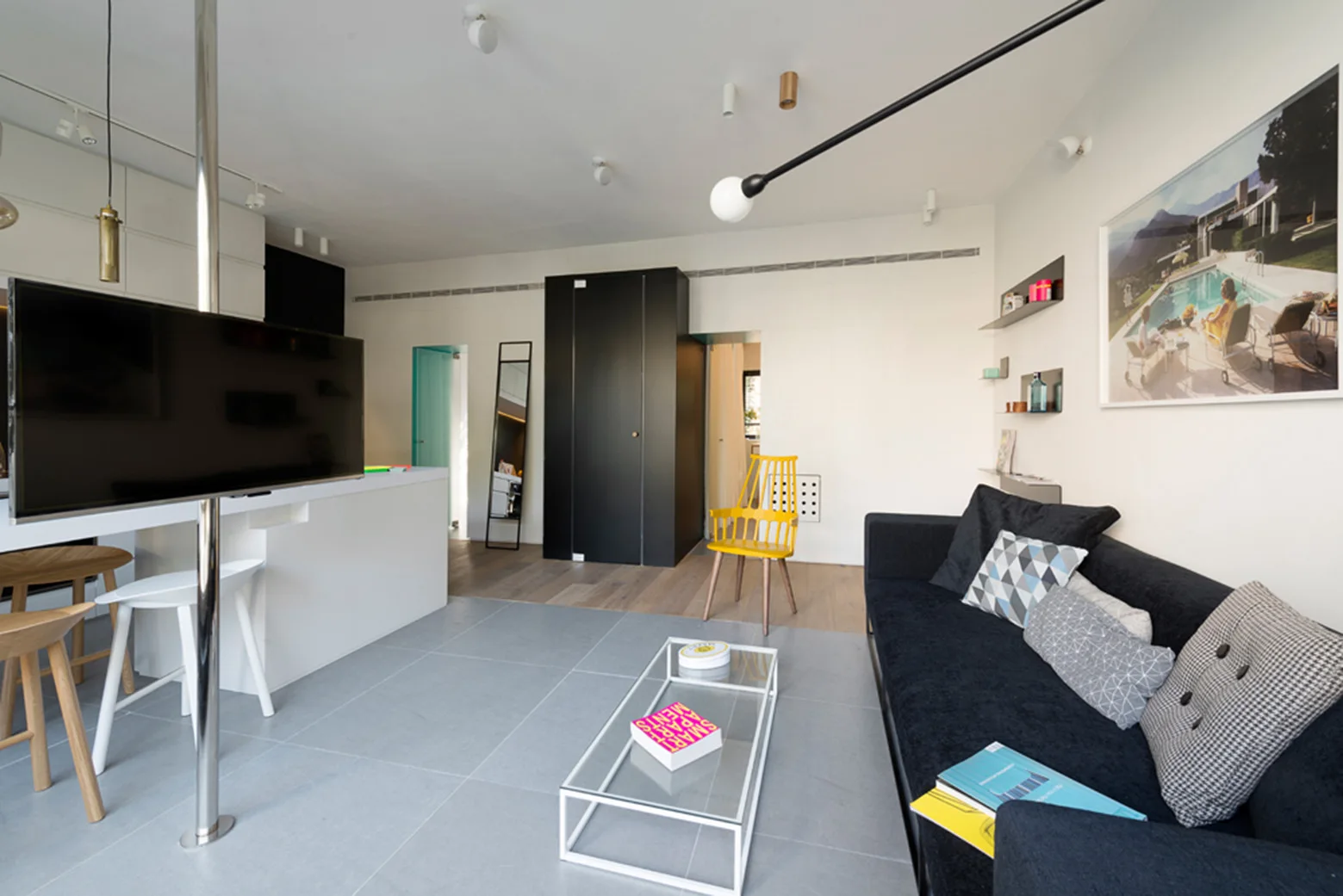 A modern open-plan living room with a black sofa, a glass-top coffee table, a yellow wood chair, and a television mounted on a metal pole.