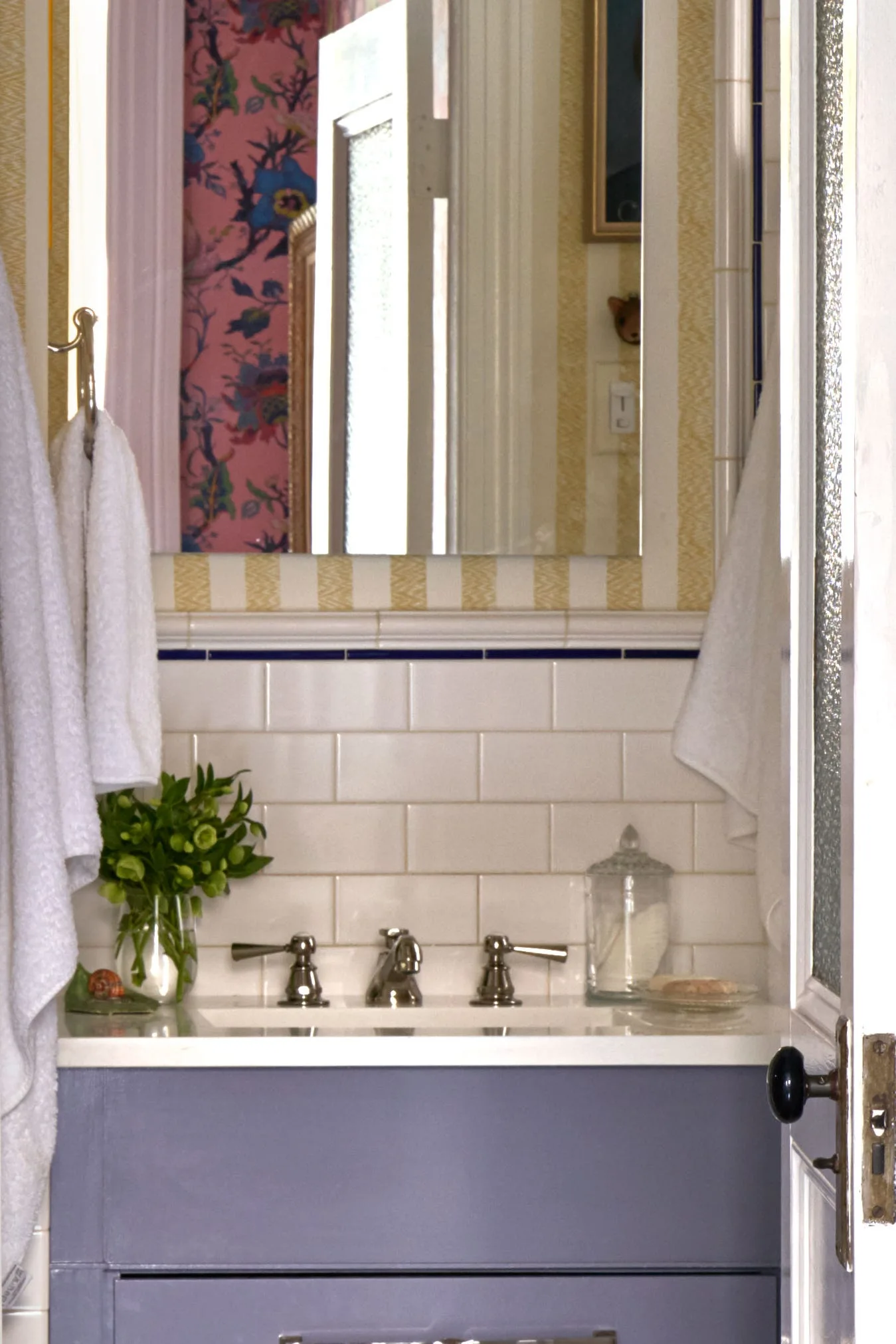 A bathroom vanity with a purple cabinet, white subway tile backsplash, and striped wallpaper beneath a rectangular mirror.