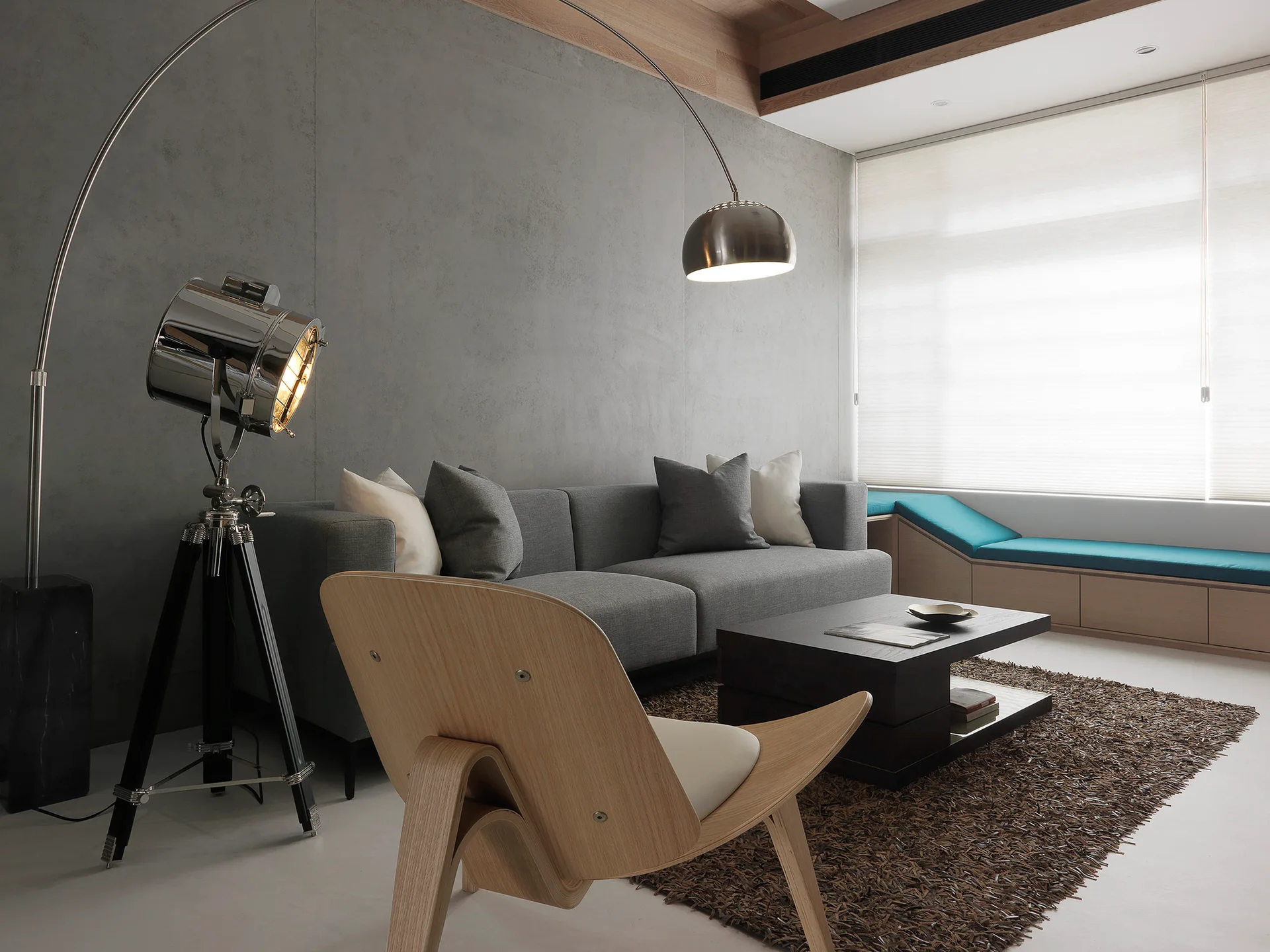 A modern living room features a gray sofa, a curved wood lounge chair, a tall metal arc lamp, and a built-in window bench.