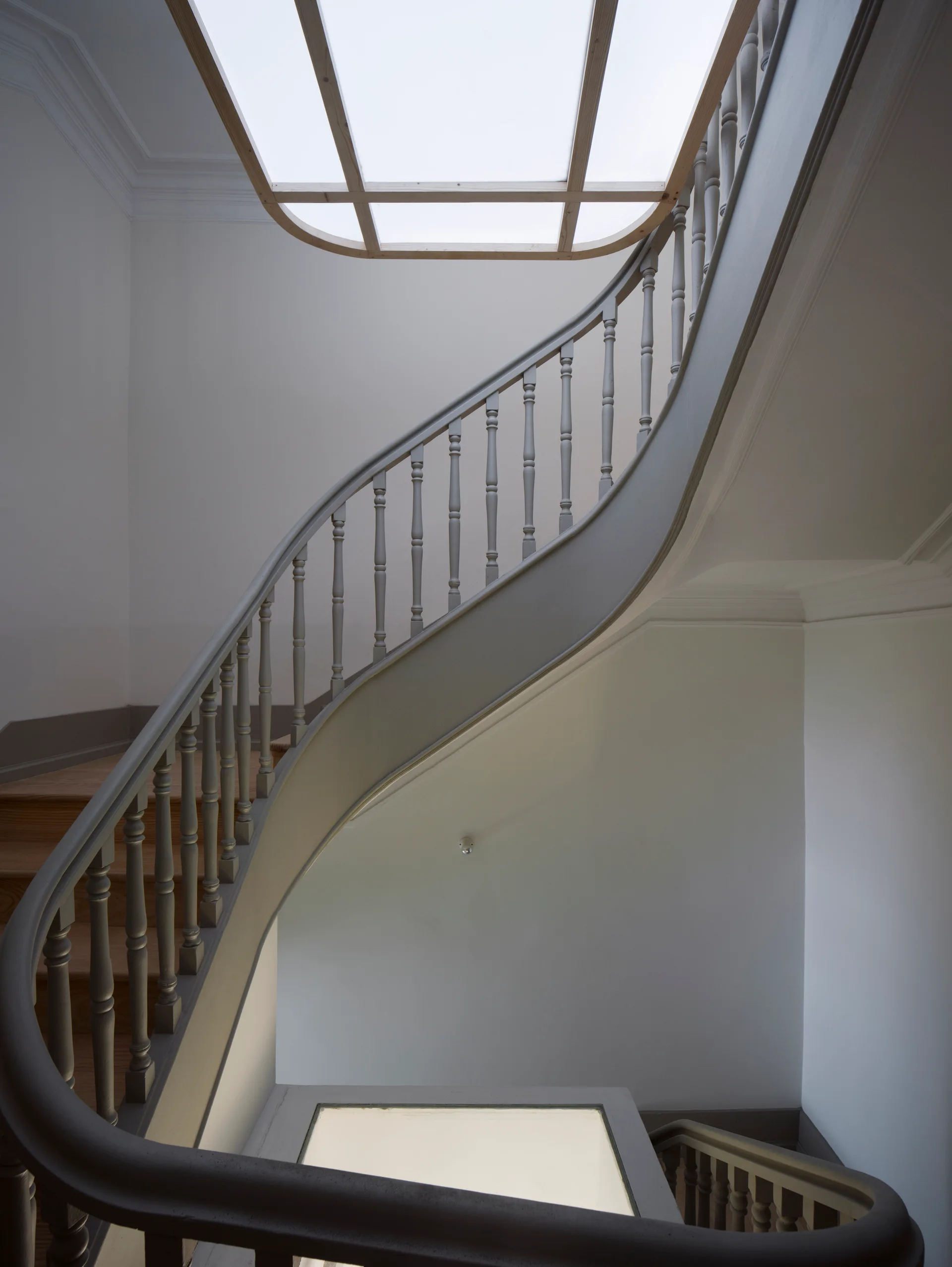 A curved wooden staircase painted grey with light wood steps ascends toward a rectangular skylight in a white-walled hall.