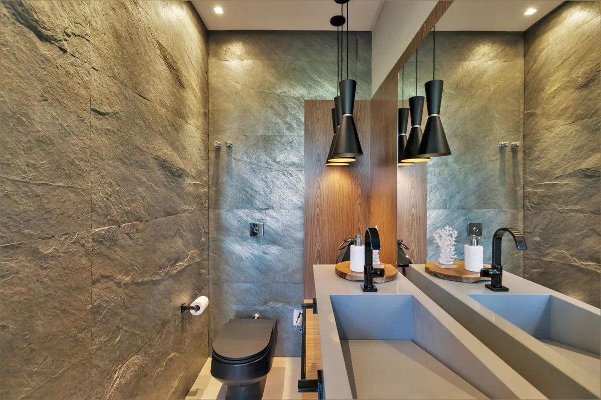 A modern bathroom features textured grey stone walls, a matte black toilet, an integrated grey sink, and black cone pendant lights.