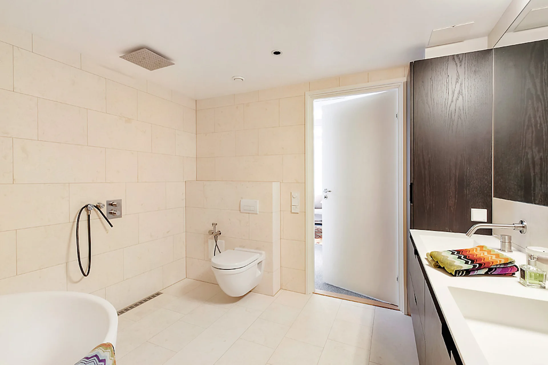 A modern bathroom features beige wall tiles, a dark wood double vanity, and a white wall-mounted toilet.