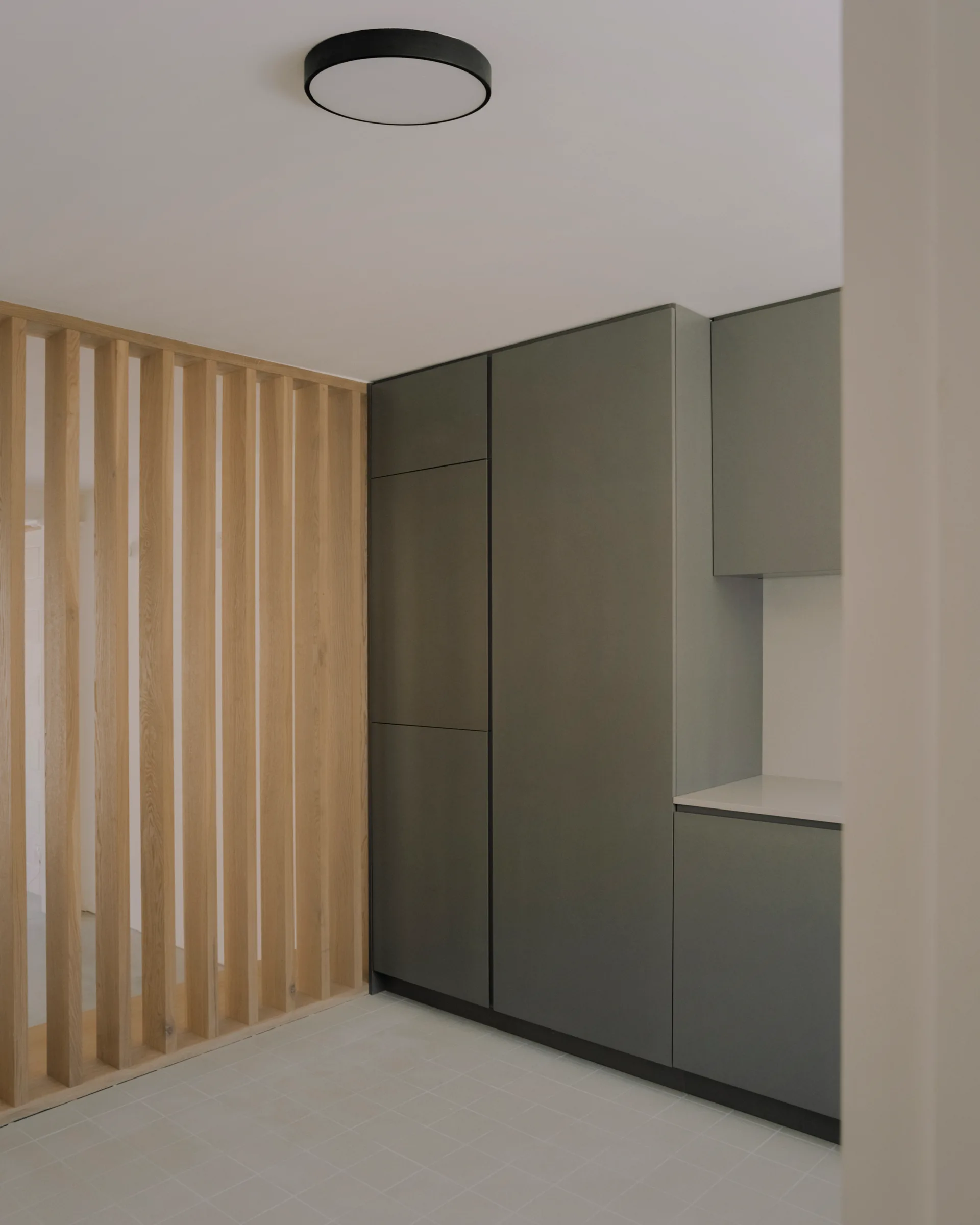 A modern kitchen entryway featuring floor-to-ceiling matte grey cabinets next to a light oak wood slatted divider screen and a tiled floor.