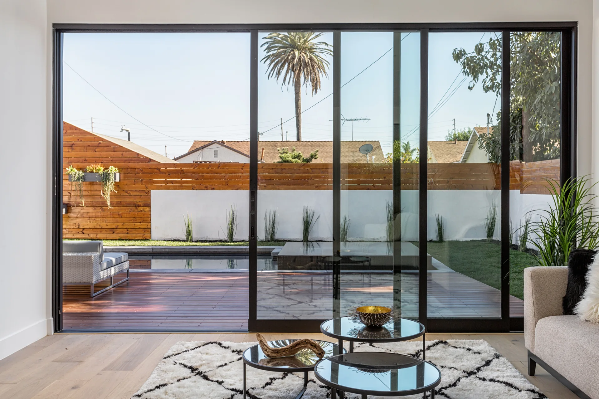 A modern living room with nesting glass coffee tables opens through large sliding glass doors to a backyard swimming pool and wooden deck.