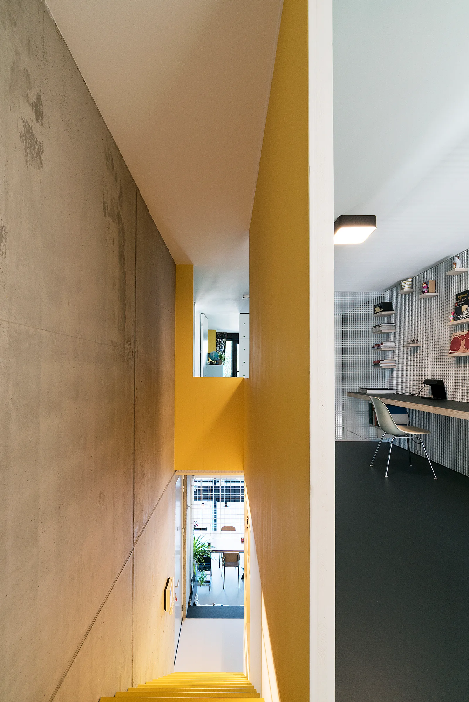 A modern home office featuring a long built-in desk and grid wall paneling adjacent to a concrete stairwell with bright yellow walls.