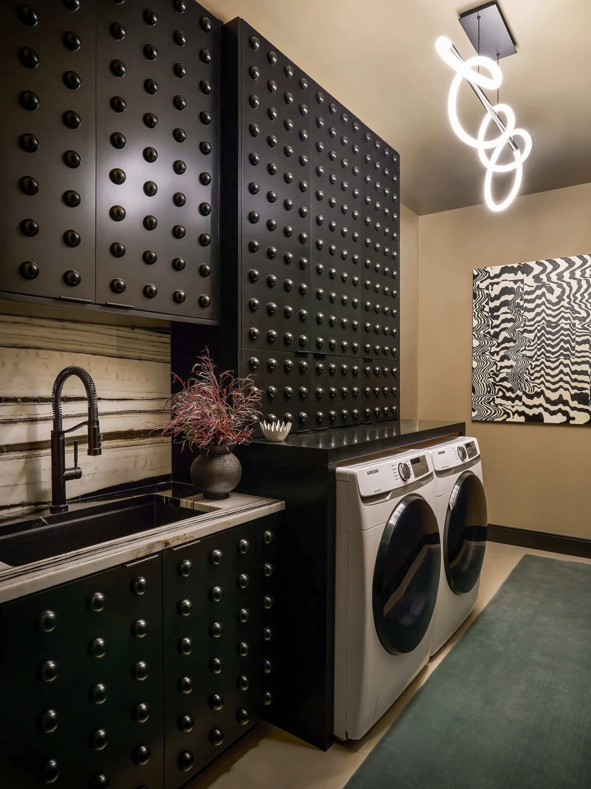 A modern laundry room features black studded cabinetry, white washing machines, a black gooseneck faucet, veined marble, and a loopy LED pendant light.