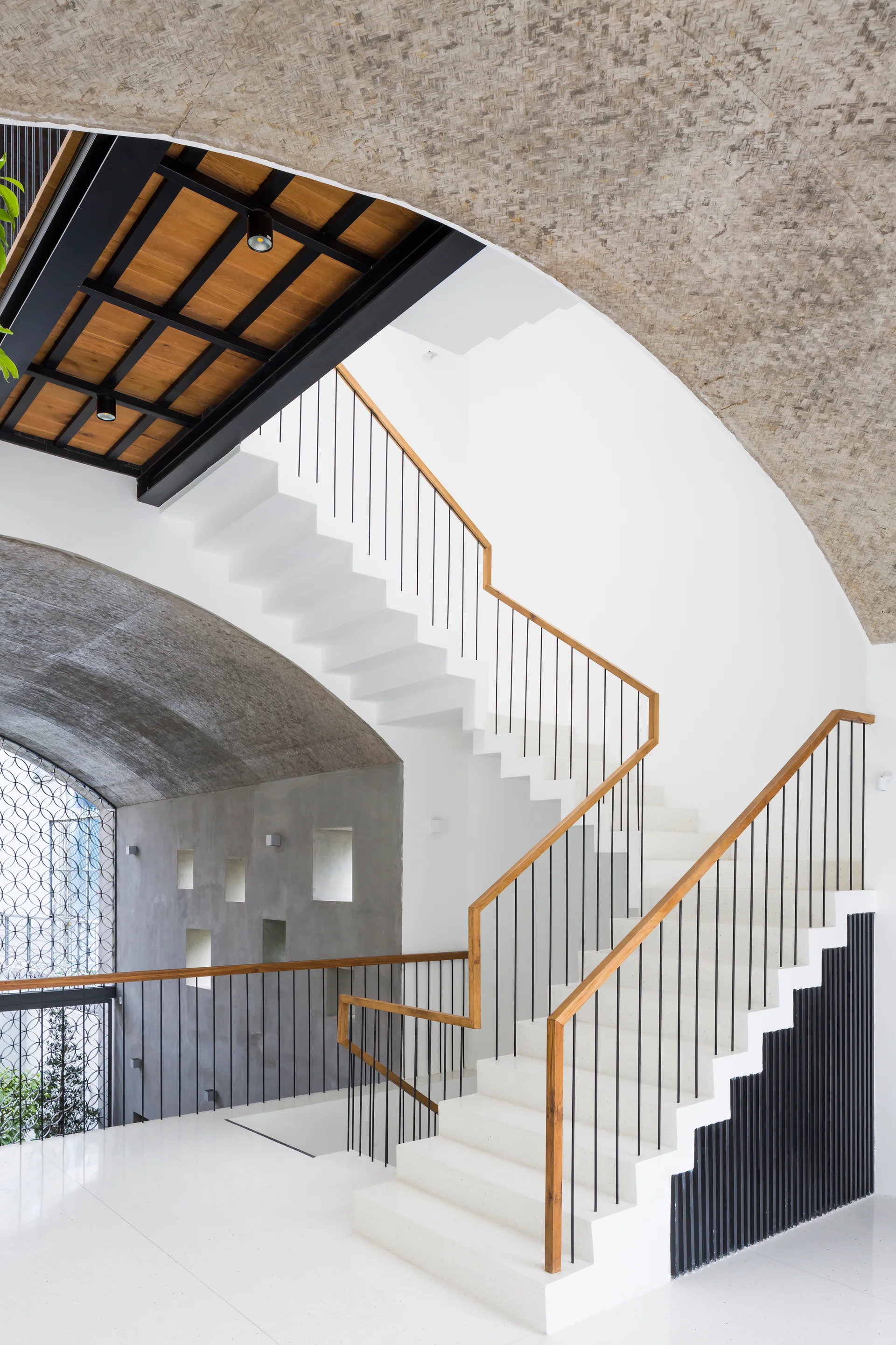 A modern white staircase featuring a warm wood handrail and black metal balusters rises beneath a curved, vaulted concrete ceiling.