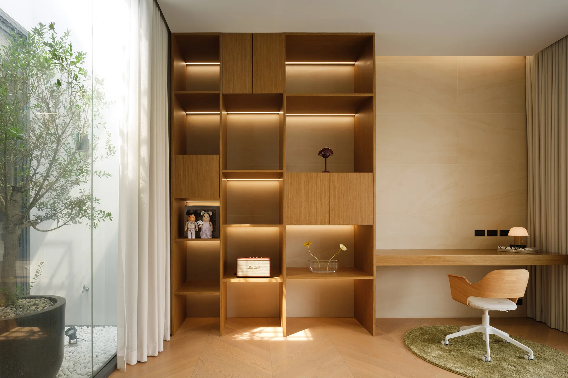 A minimalist home office features a large wooden built-in shelving unit, a matching desk, a round green rug under an office chair, and a glass wall.