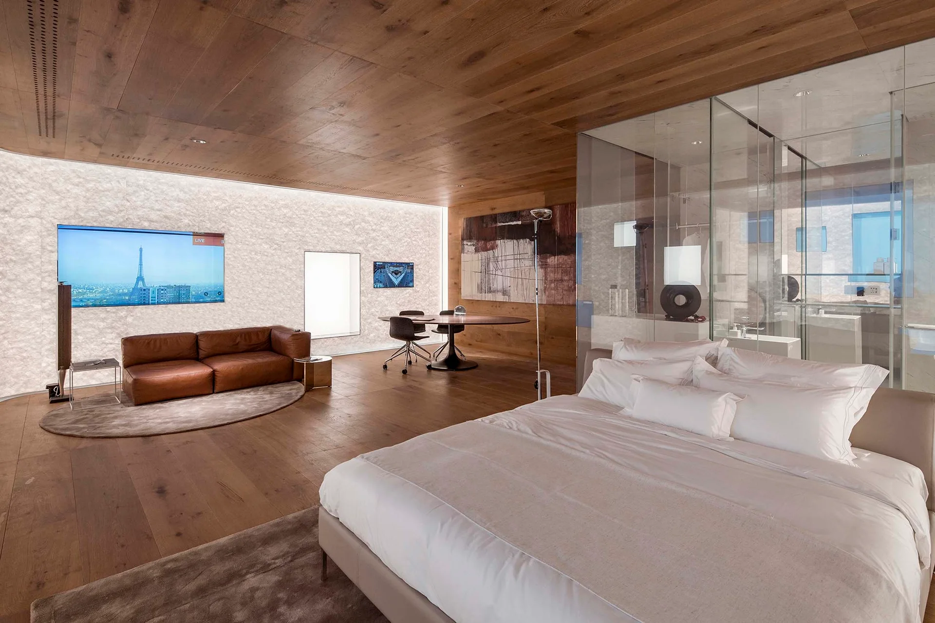 A spacious bedroom features matching wood panels on the ceiling and floor, a glass-enclosed bathroom, and a leather sofa seating area.