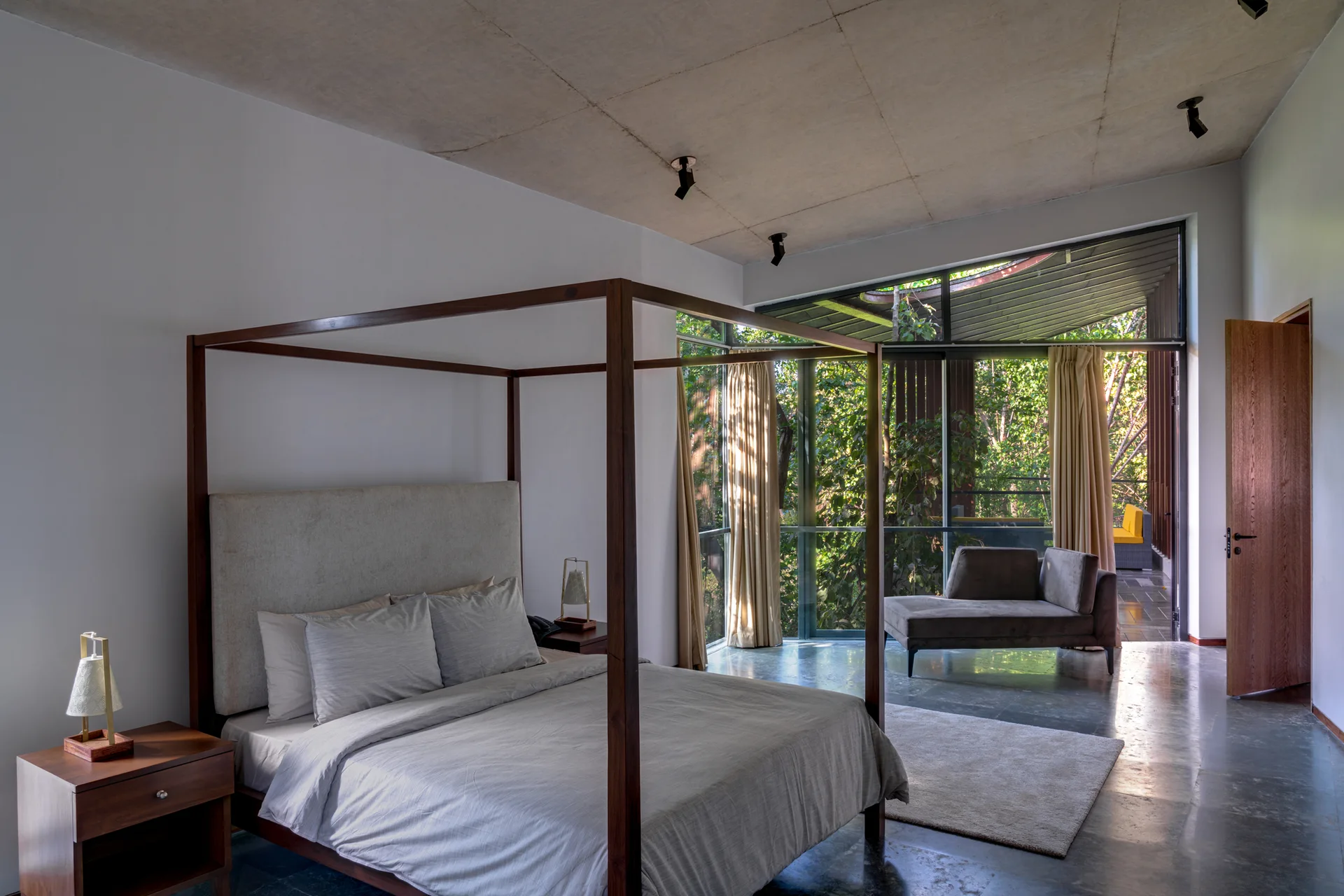 A modern bedroom features a wooden four-poster bed, an exposed concrete ceiling, and large floor-to-ceiling windows facing green trees.