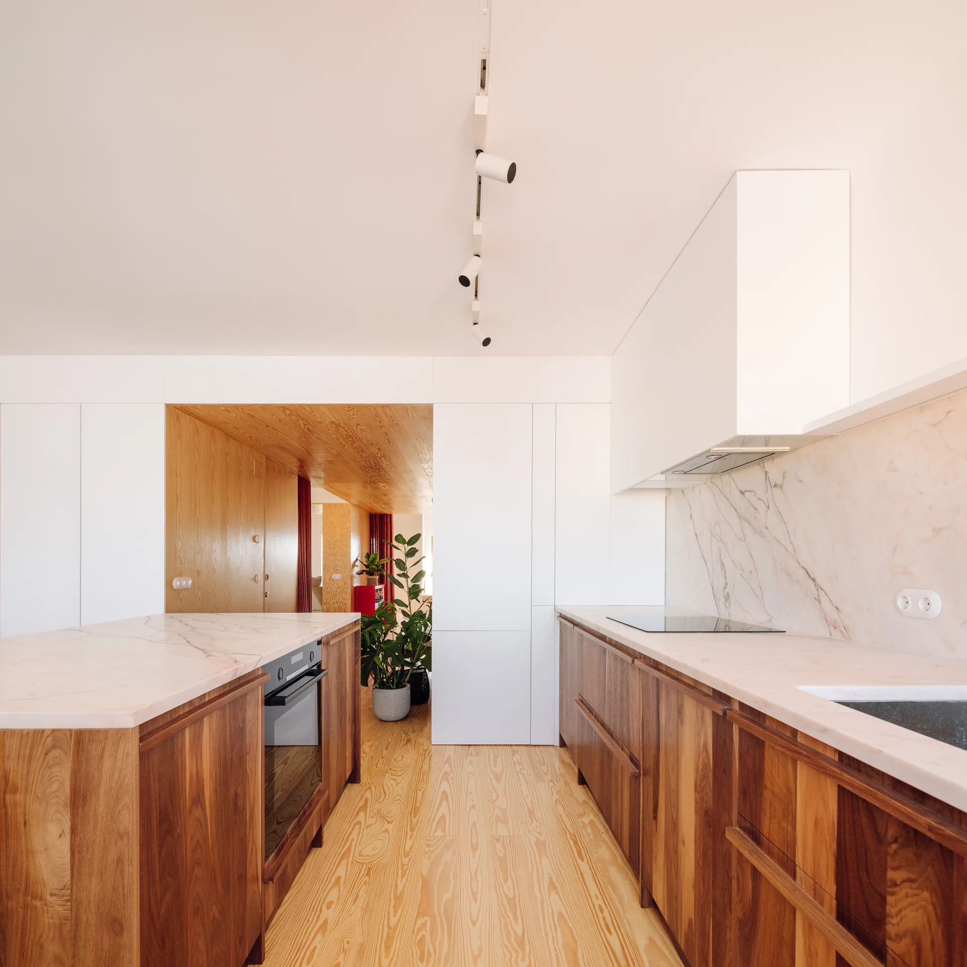 A modern kitchen featuring rich wood cabinets, pinkish veined marble countertops, light wood floors, and minimalist white upper cabinetry.