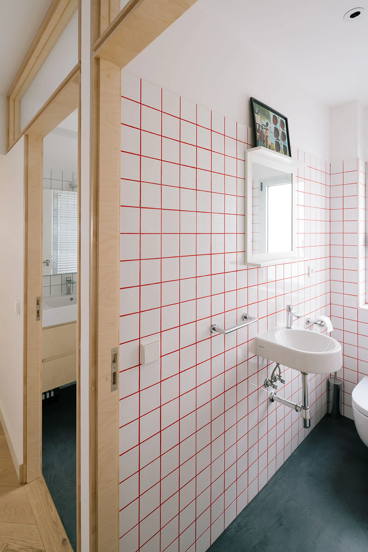 A modern bathroom features white tiled walls with contrast red grout, a wall-mounted sink, dark floors, and a light wood door frame.
