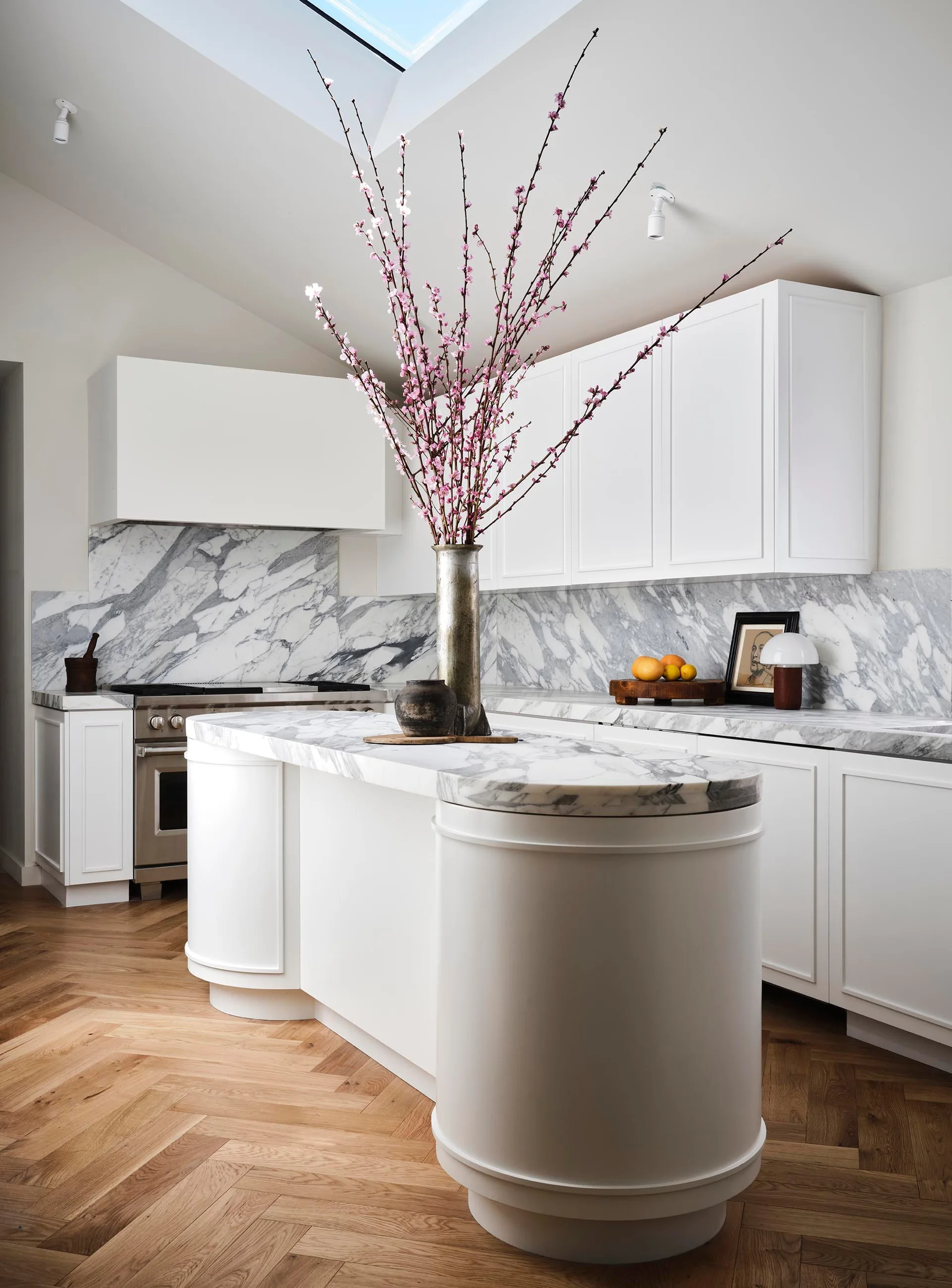 A bright kitchen features a curved white island with a veined marble countertop, white cabinetry, a marble backsplash, and herringbone wood floors.