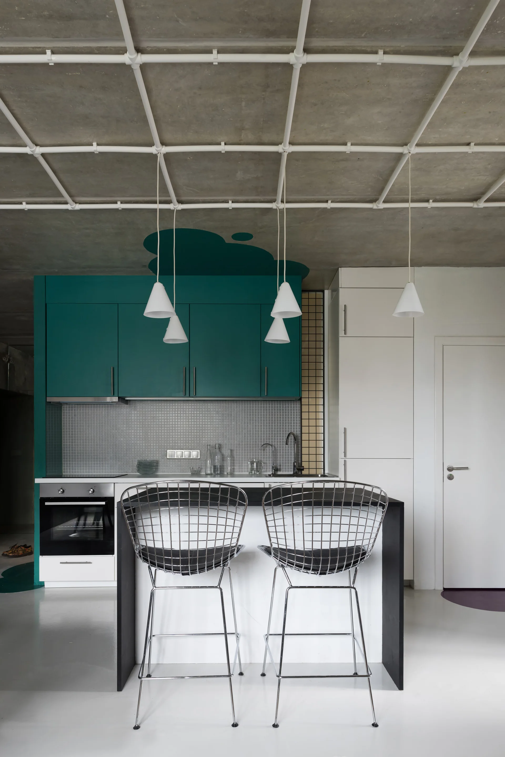 A modern industrial kitchen with teal and white cabinetry, a concrete ceiling with exposed pipes, and two metal wire bar stools.