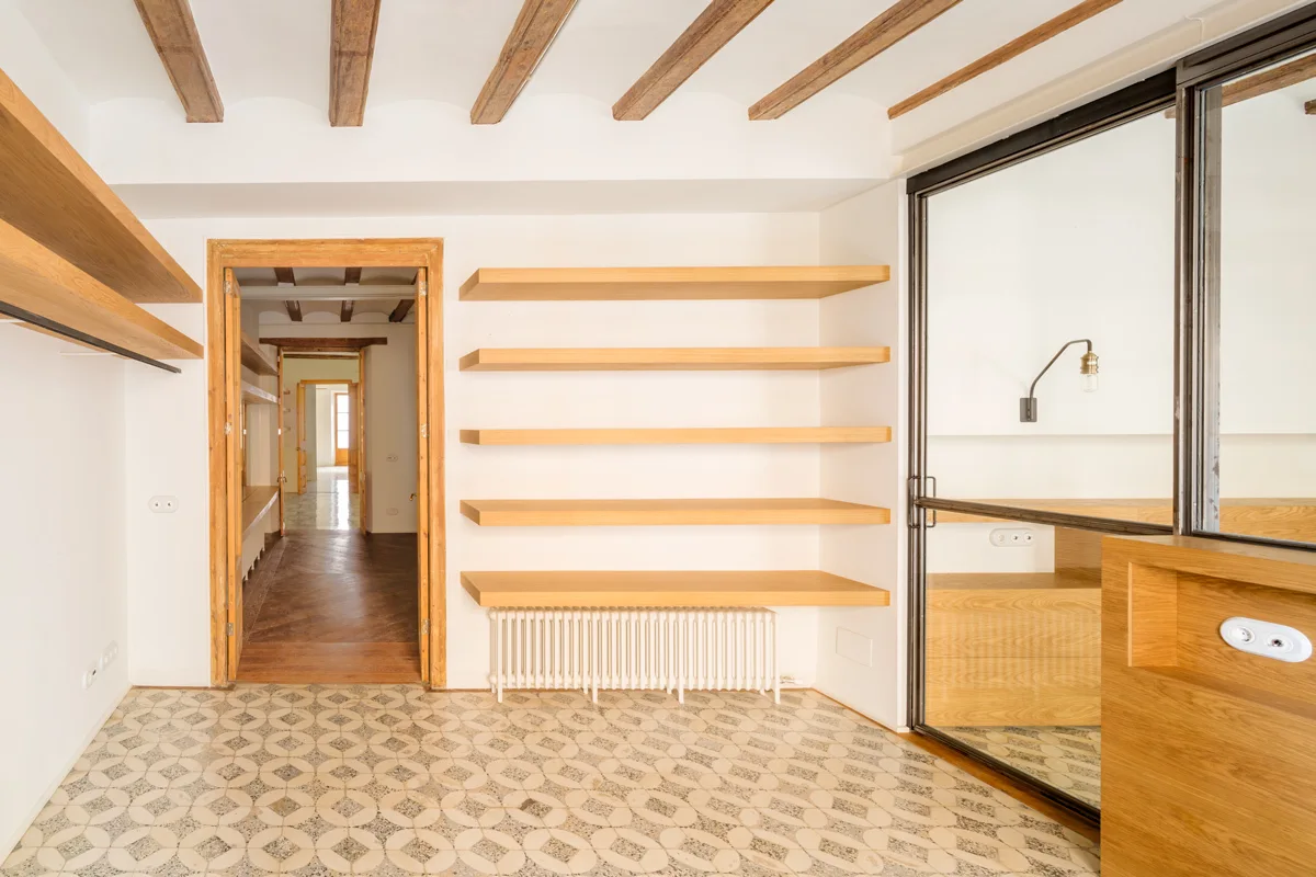 A bright interior hallway features patterned tile flooring, exposed wooden ceiling beams, and integrated light-wood shelving above a white radiator.