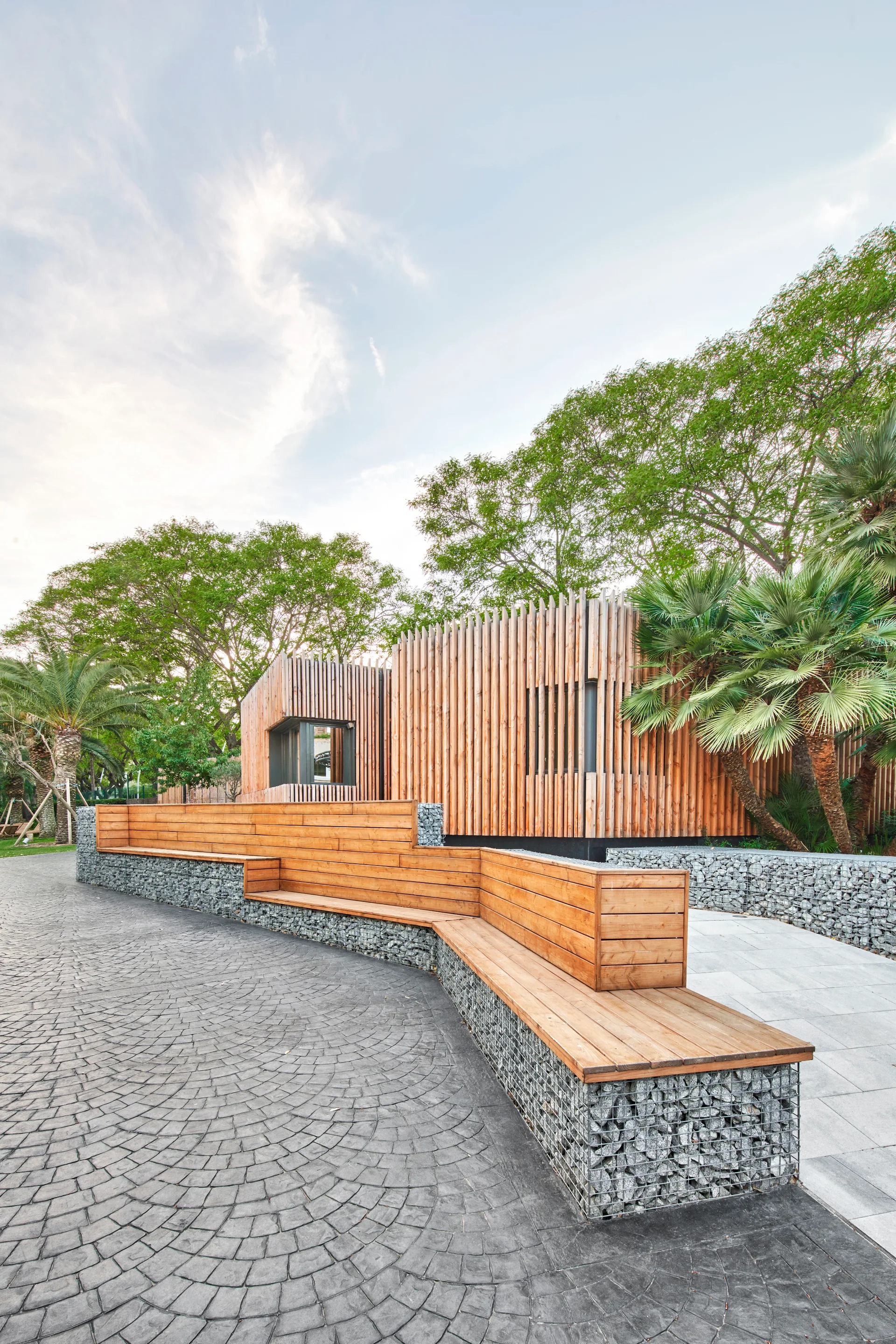 A tiered wood bench sits on stone gabion walls alongside a courtyard paved with gray stone, with a wood-slatted building in the background.