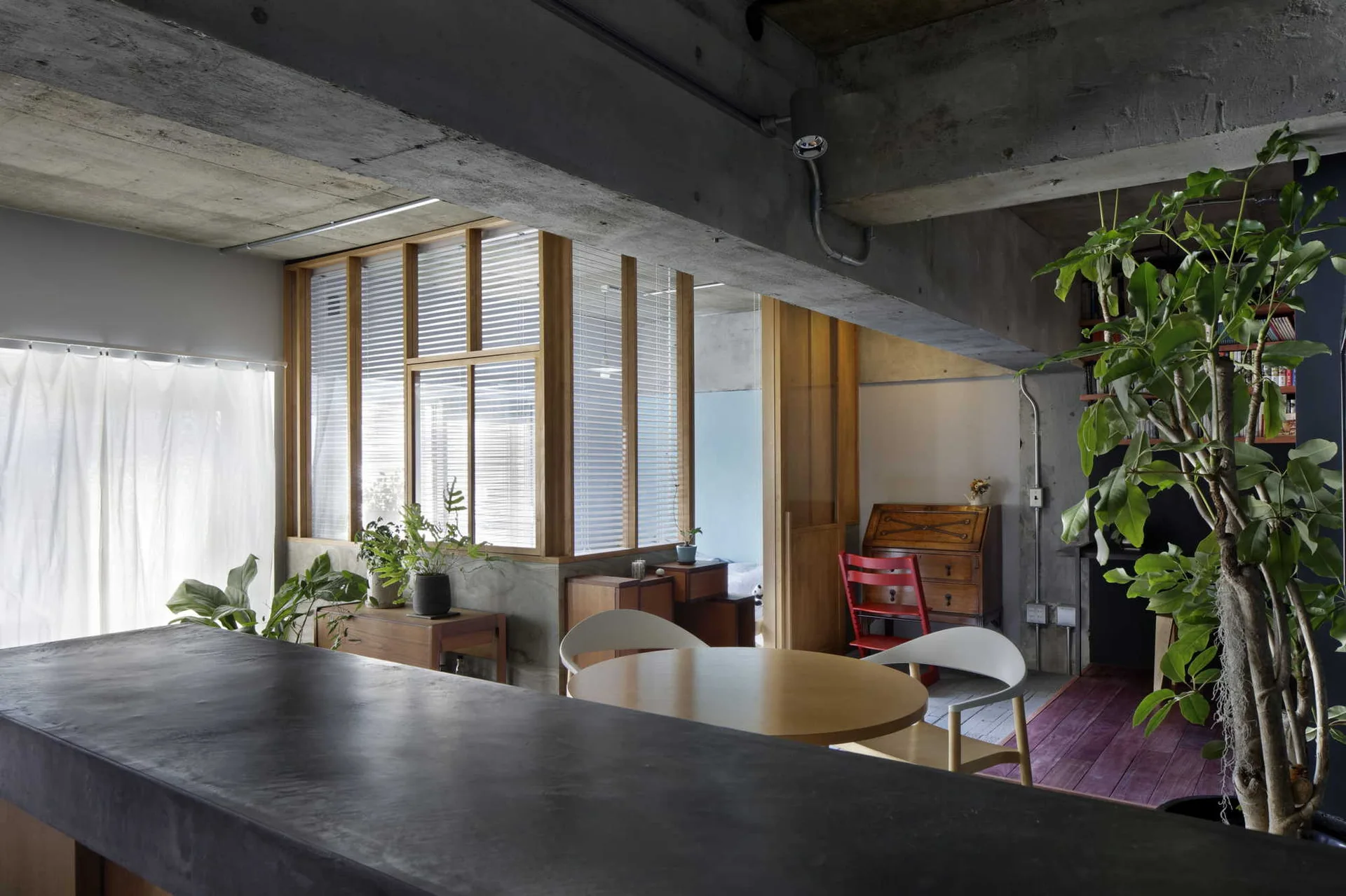 An industrial dining area features an exposed concrete ceiling, a round wooden table with white chairs, and an interior wood-framed partition wall.