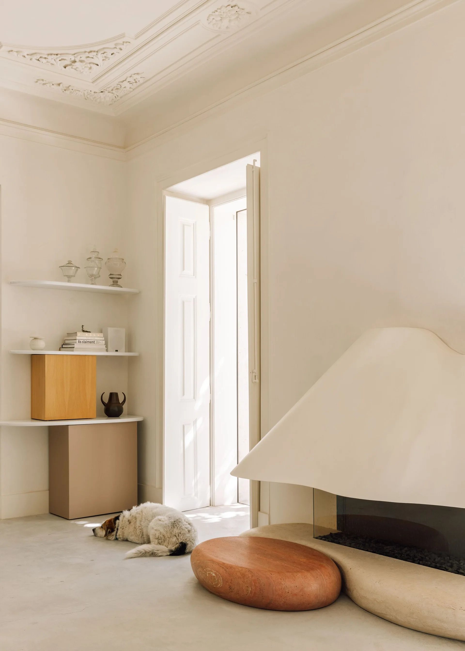 A minimalist cream living room features a curved sculptural fireplace, asymmetrical corner shelving with wood cabinets, and ornate ceiling moldings.