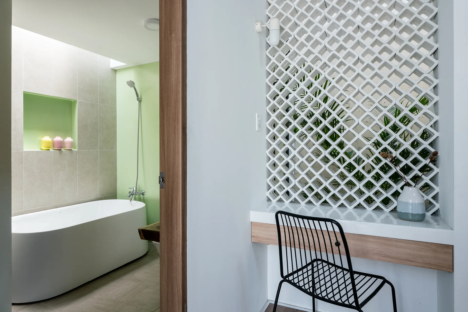 A view through a wood door frame showing a white freestanding tub against a light green wall and a white geometric breeze block screen above a floating desk.