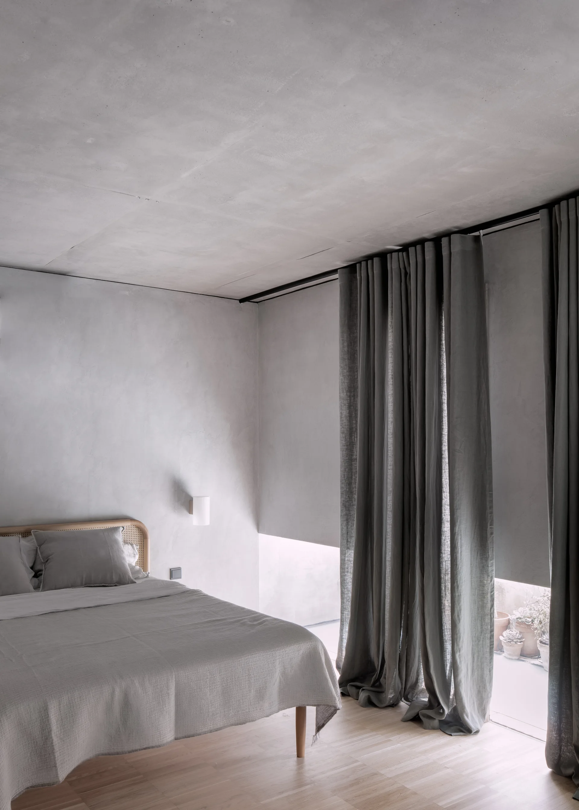 A minimalist bedroom features a bed with a cane headboard, grey linens, and tall charcoal curtains against a textured concrete wall.