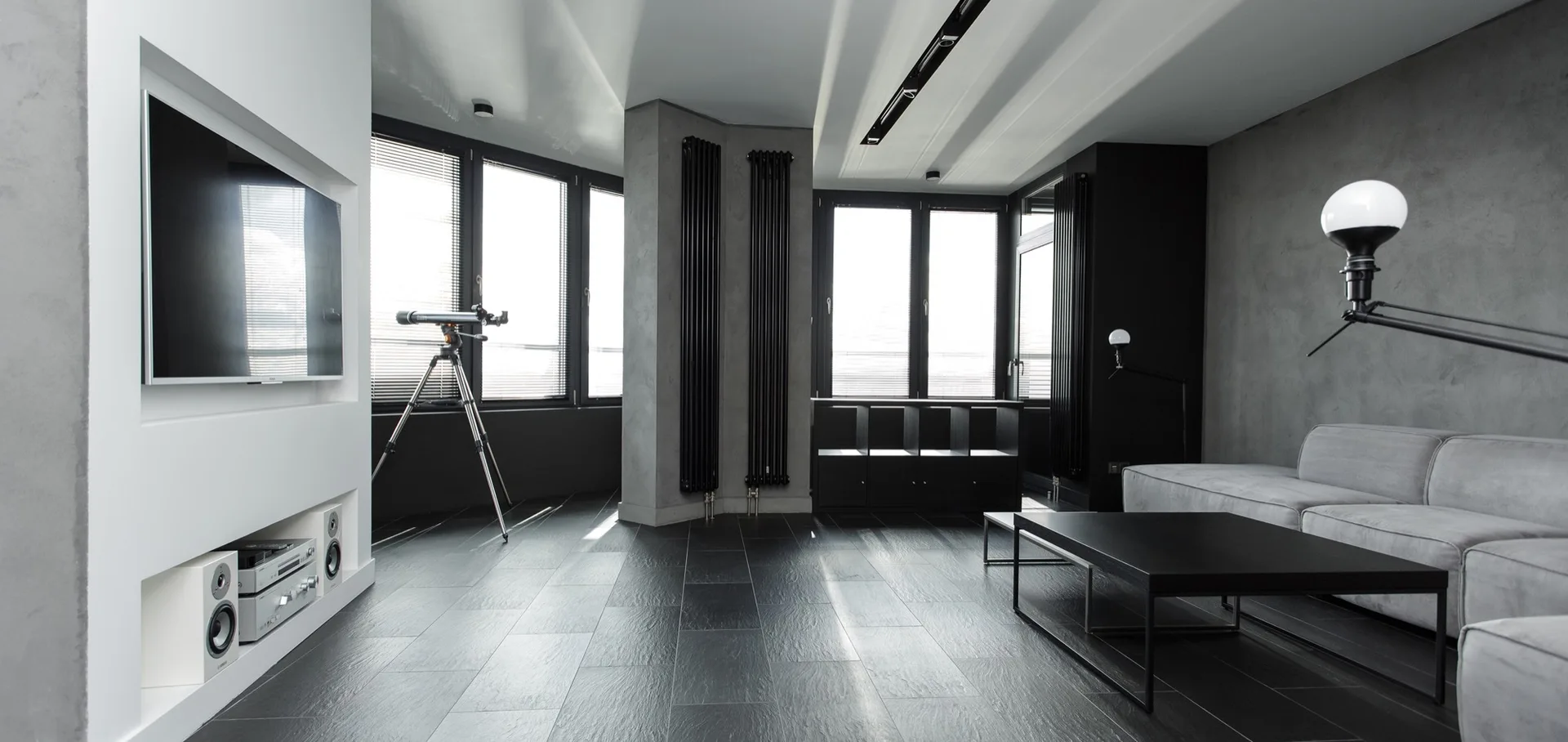 A minimalist modern living room with a gray sofa, slate tile floors, a white built-in media wall, and tall black vertical radiators.
