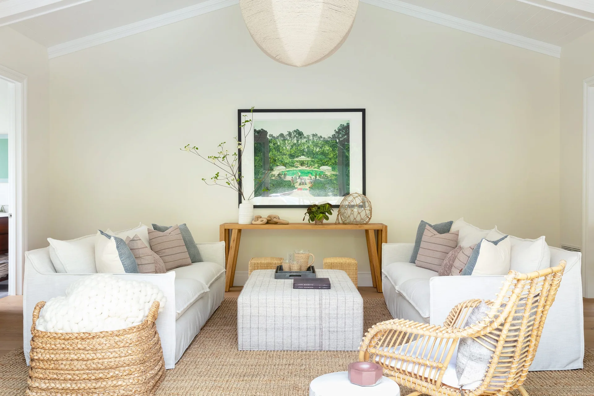 Symmetrical living room with two white sofas facing a plaid upholstered ottoman, a wooden console table with framed art, and a rattan chair.