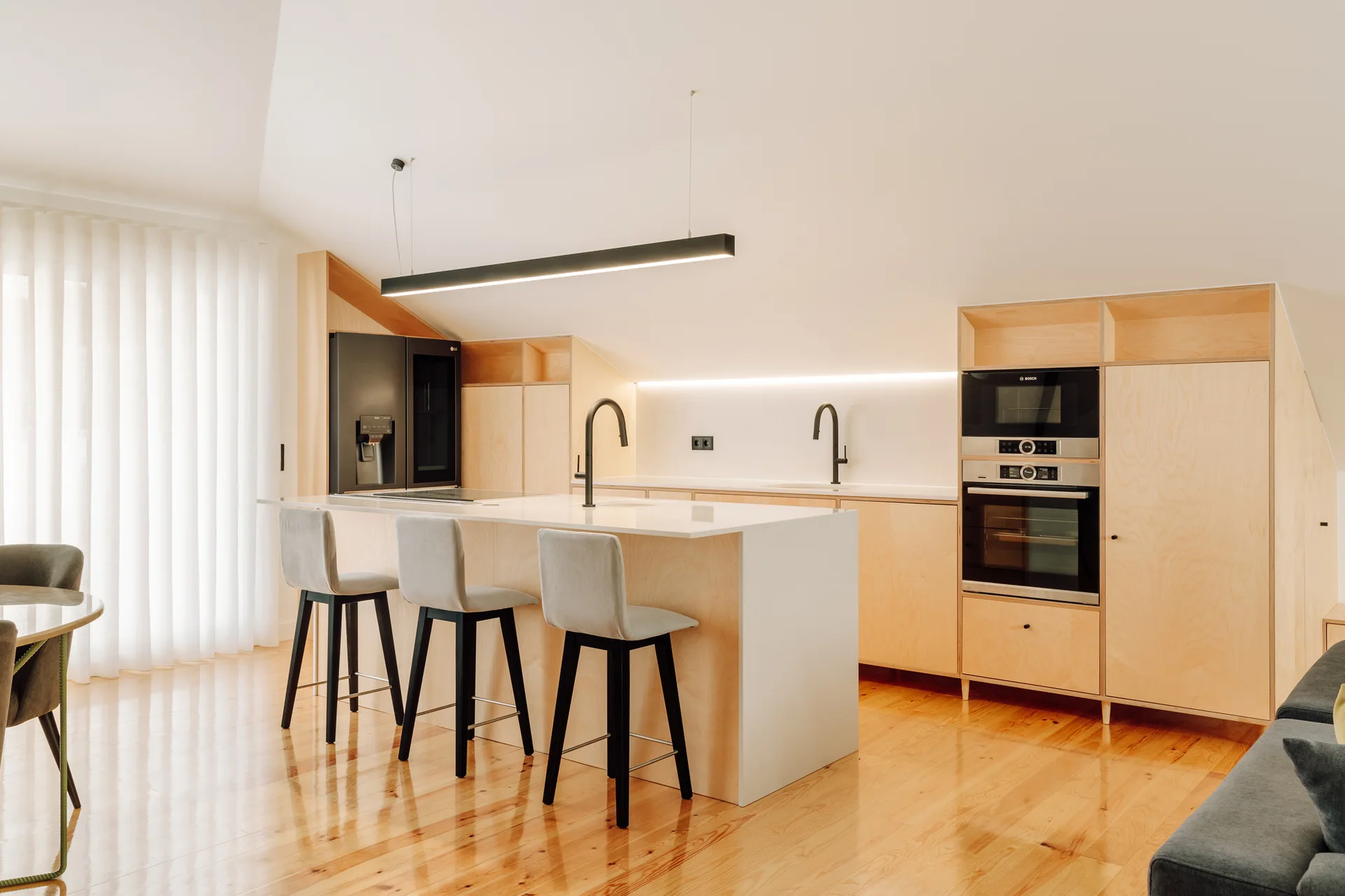 A modern minimalist kitchen features light plywood cabinetry custom-built to fit a sloped ceiling, a white kitchen island, and three bar stools.