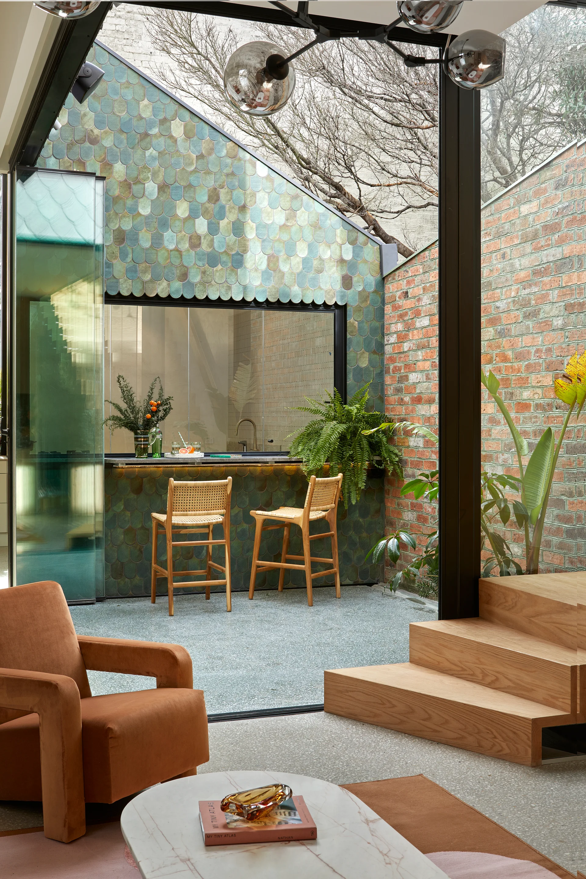 A modern living room with a rust velvet armchair overlooks a glass-walled patio featuring a teal scalloped tile bar and two wood bar stools.