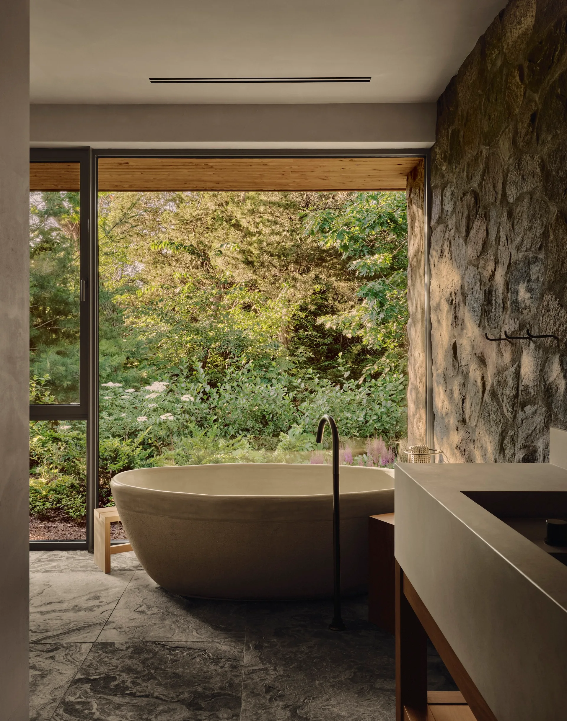 A modern bathroom features a freestanding concrete tub set against a large window overlooking trees, beside a textured stone wall.