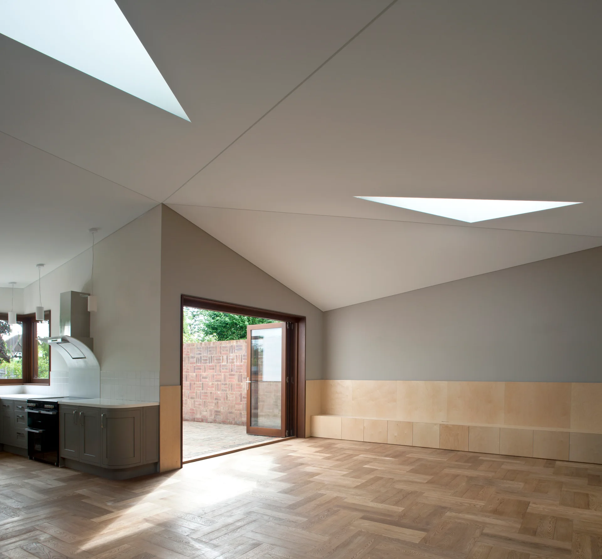 An open kitchen and living area with a sloped ceiling, triangular skylights, grey walls, a plywood bench, and herringbone wood floors.