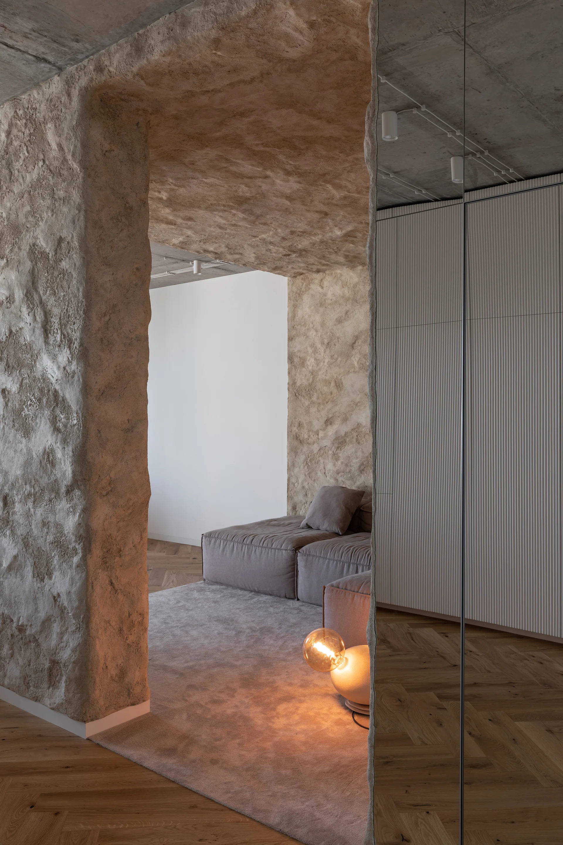 A minimalist living room features a rough-textured beige stone archway, a low-profile gray sofa, herringbone wood floors, and a glowing globe floor lamp.