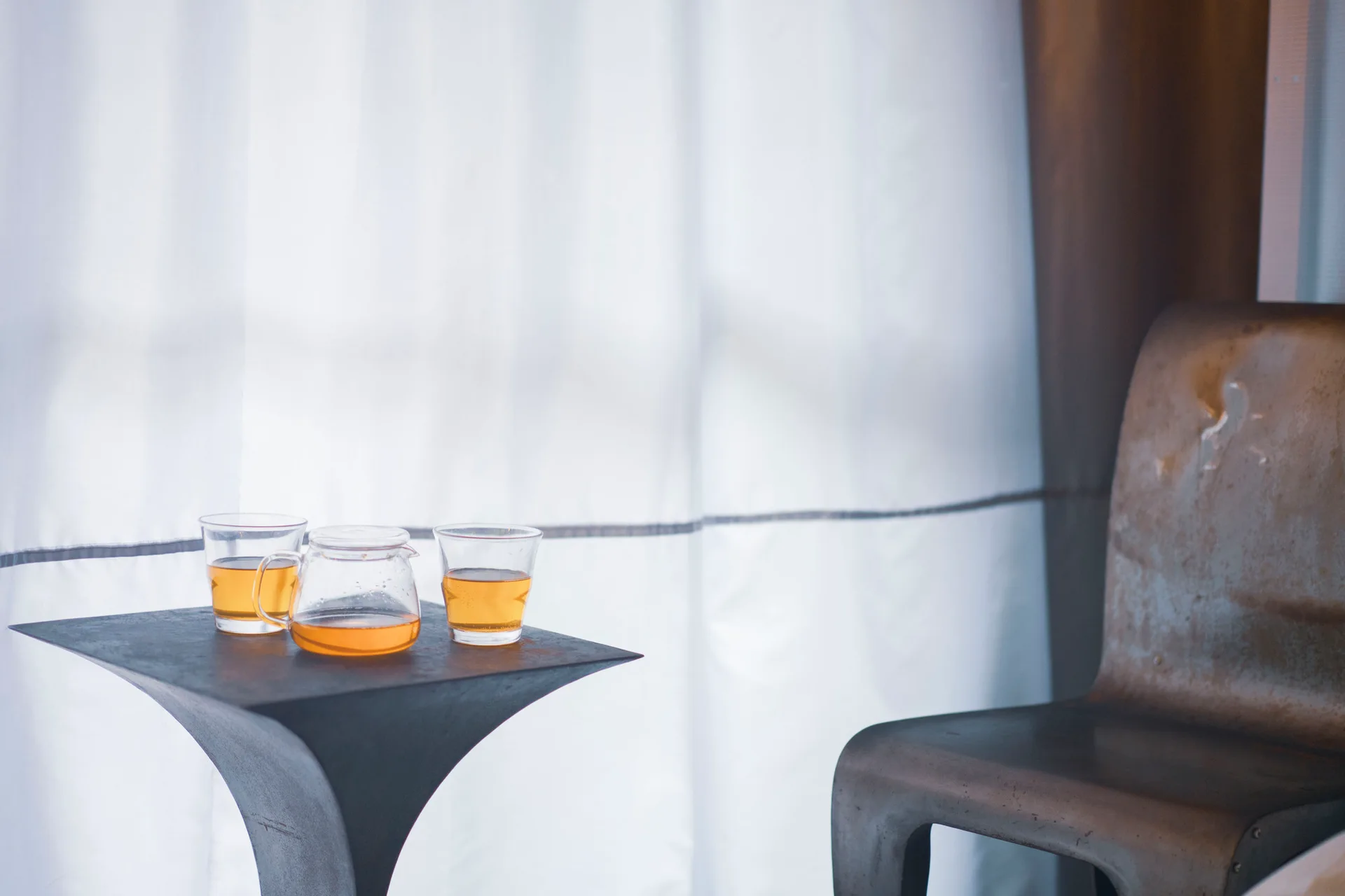 A dark sculptural metal side table holding a glass teapot and cups sits next to a distressed metal chair in front of sheer white curtains.
