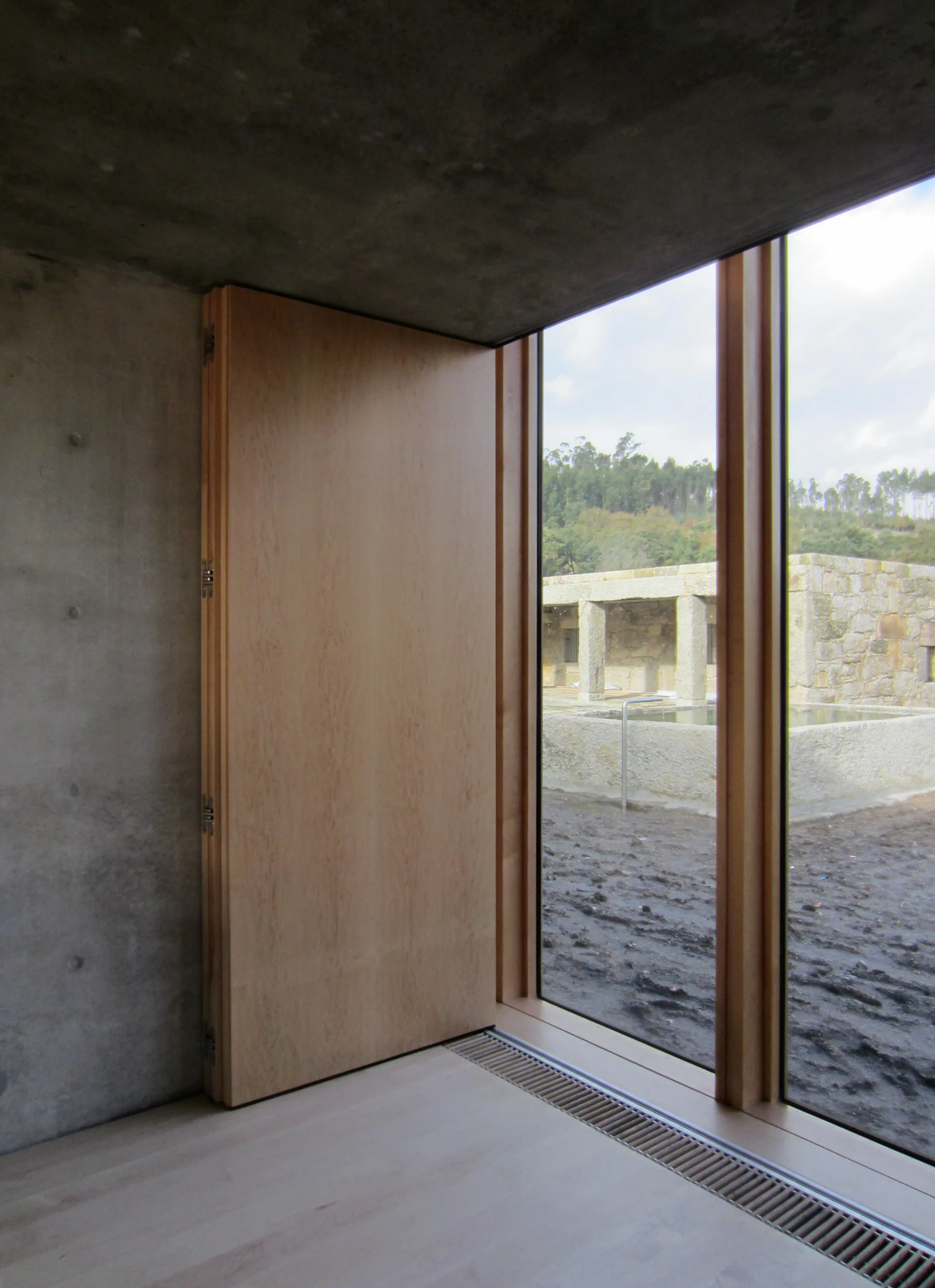 A minimalist room featuring raw concrete walls, a large light wood folding door, and a tall wood-framed window looking outside.