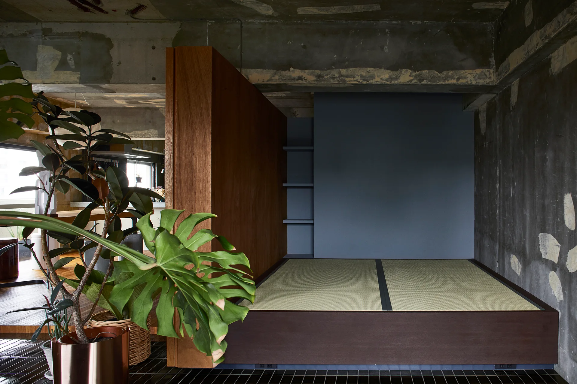 A raised platform with tatami mats sits next to a tall wood divider, set against raw concrete walls and a blue-grey painted alcove.