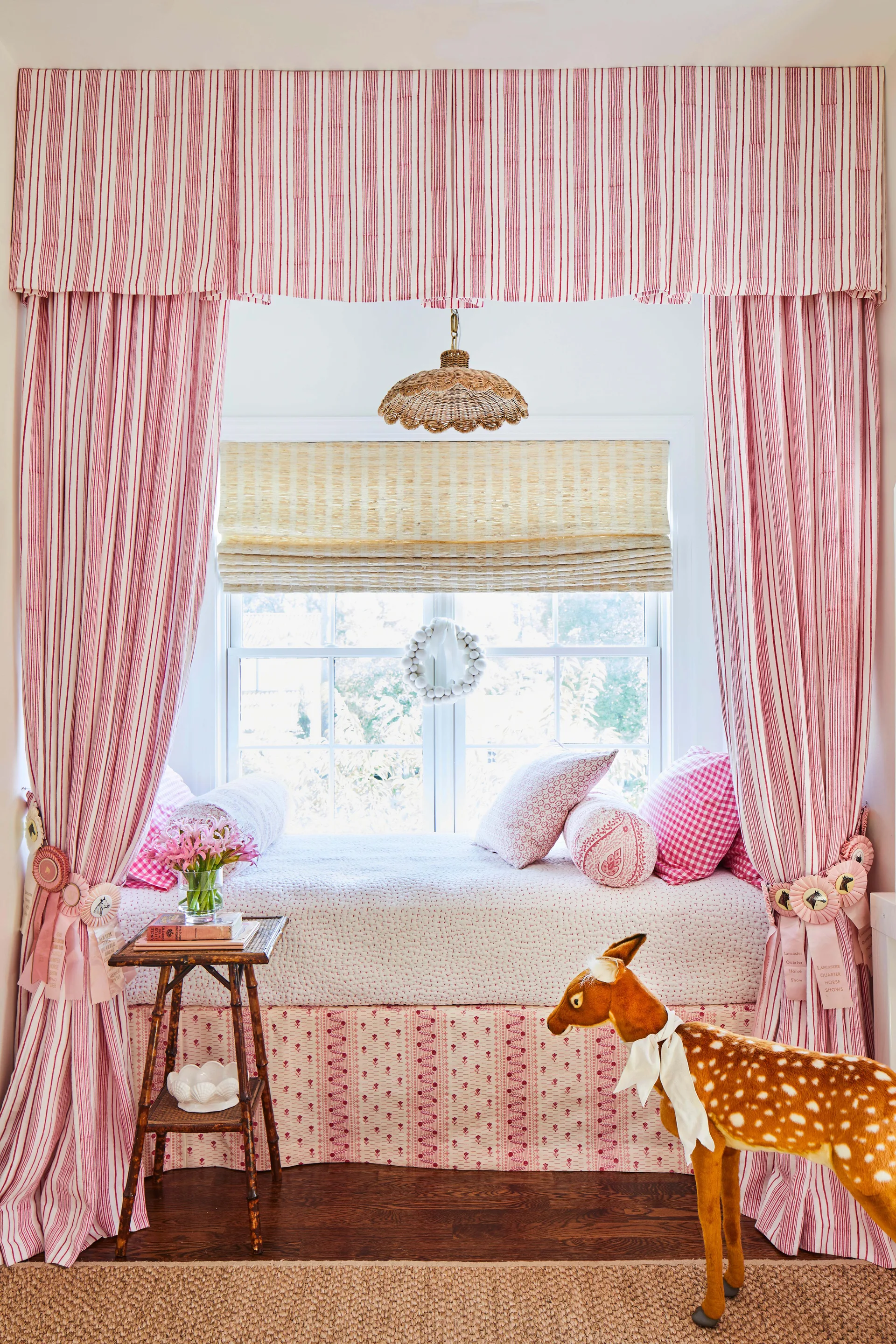 A built-in window seat with pink pillows is framed by red striped curtains and features a woven roman shade and a plush deer toy.
