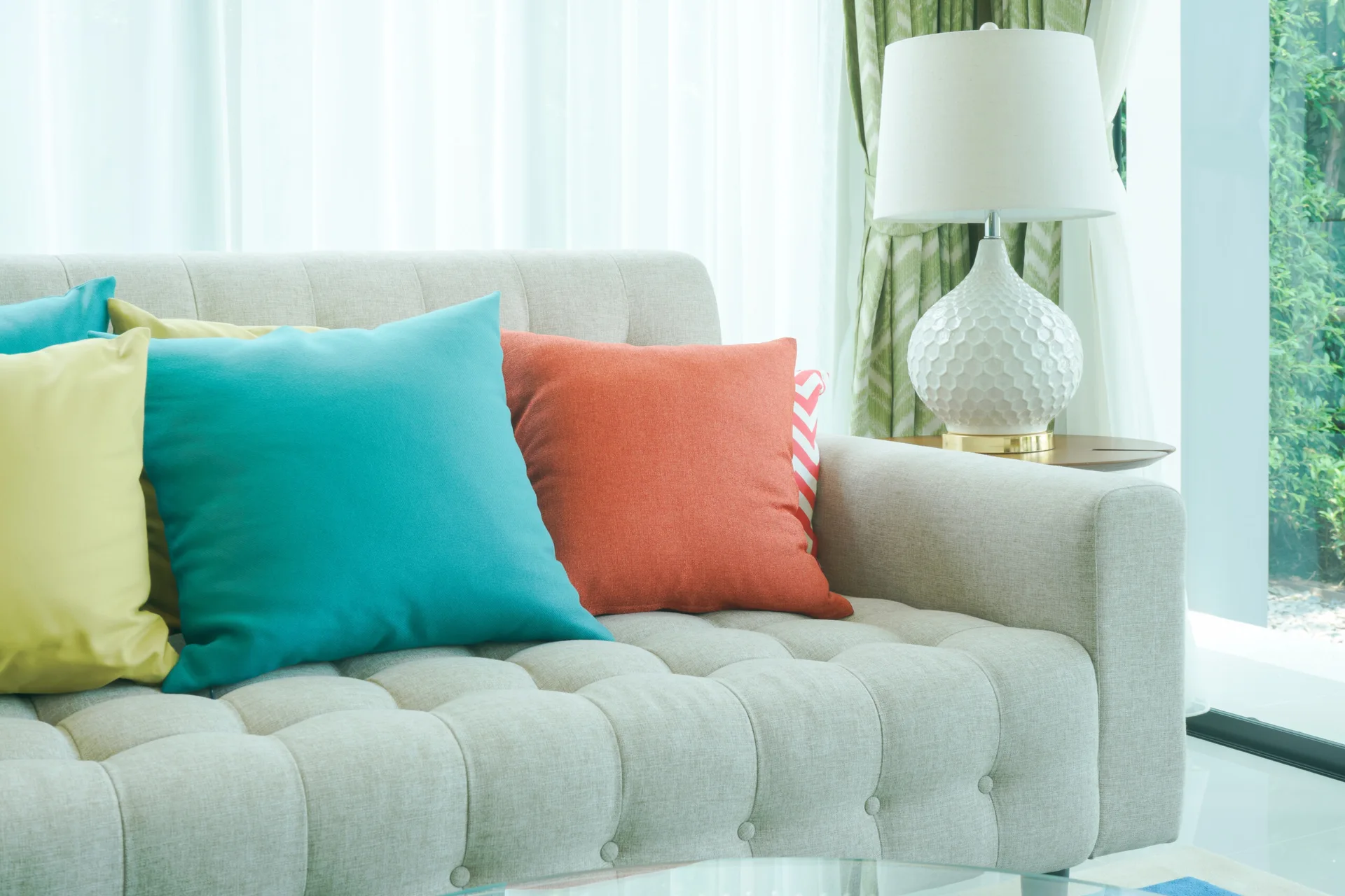 A beige button-tufted sofa decorated with colorful pillows sits next to a white ceramic lamp with a honeycomb texture.