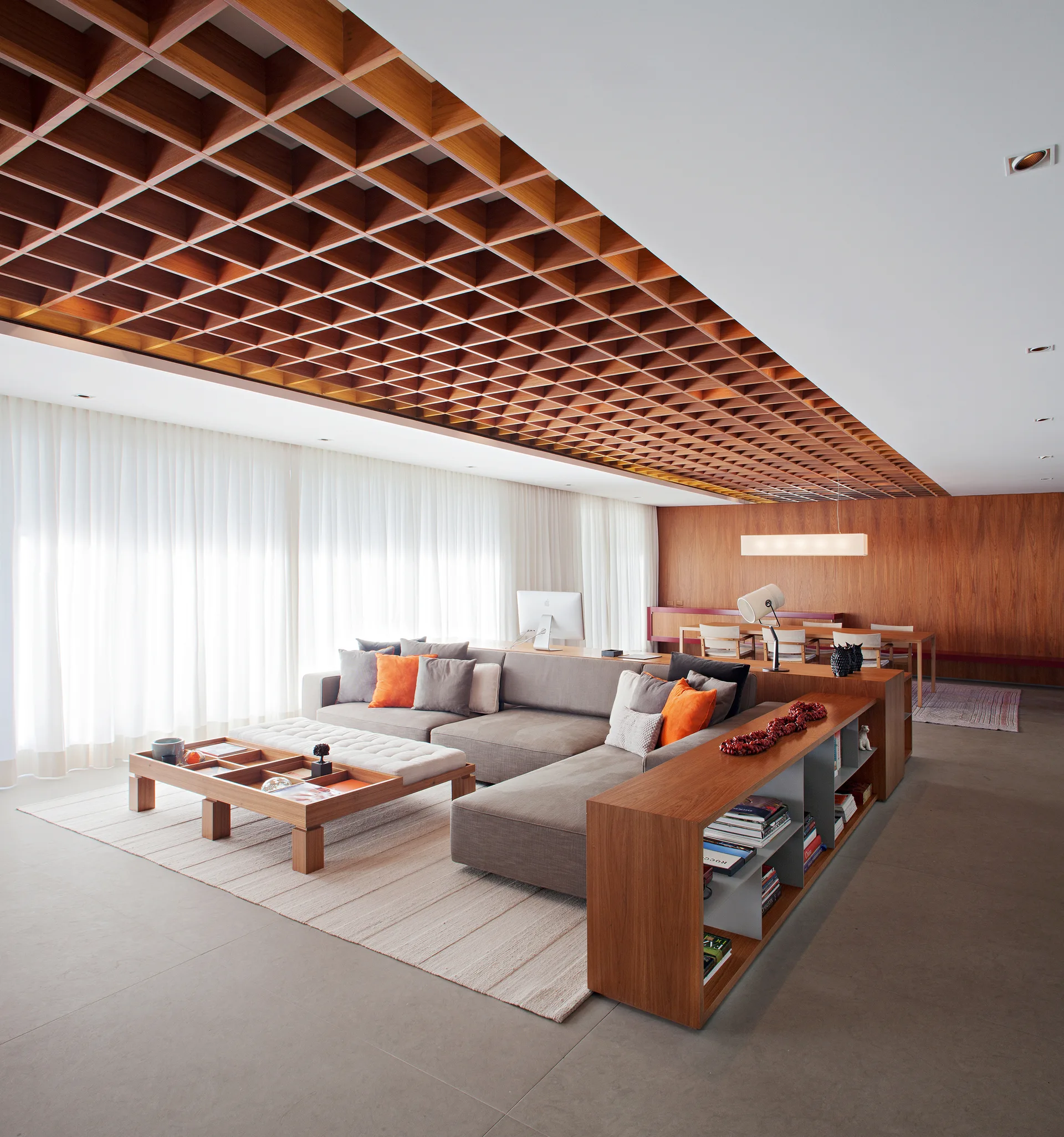 A spacious living room featuring a large grey sectional sofa, an integrated wood console shelf, a timber coffered ceiling, and sheer white curtains.